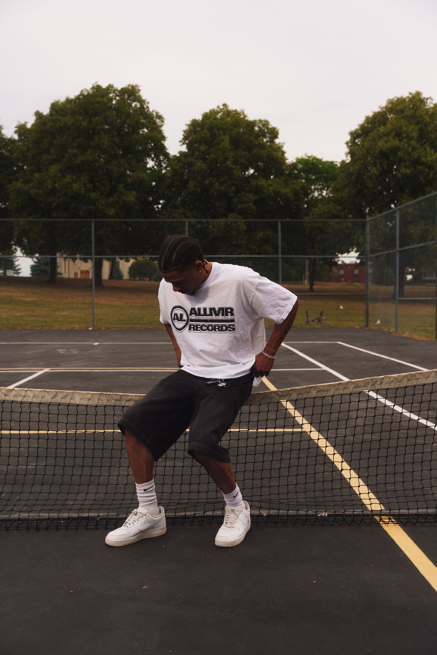 Person wearing white ALLMVR Records t-shirt and shorts sitting on tennis court net.