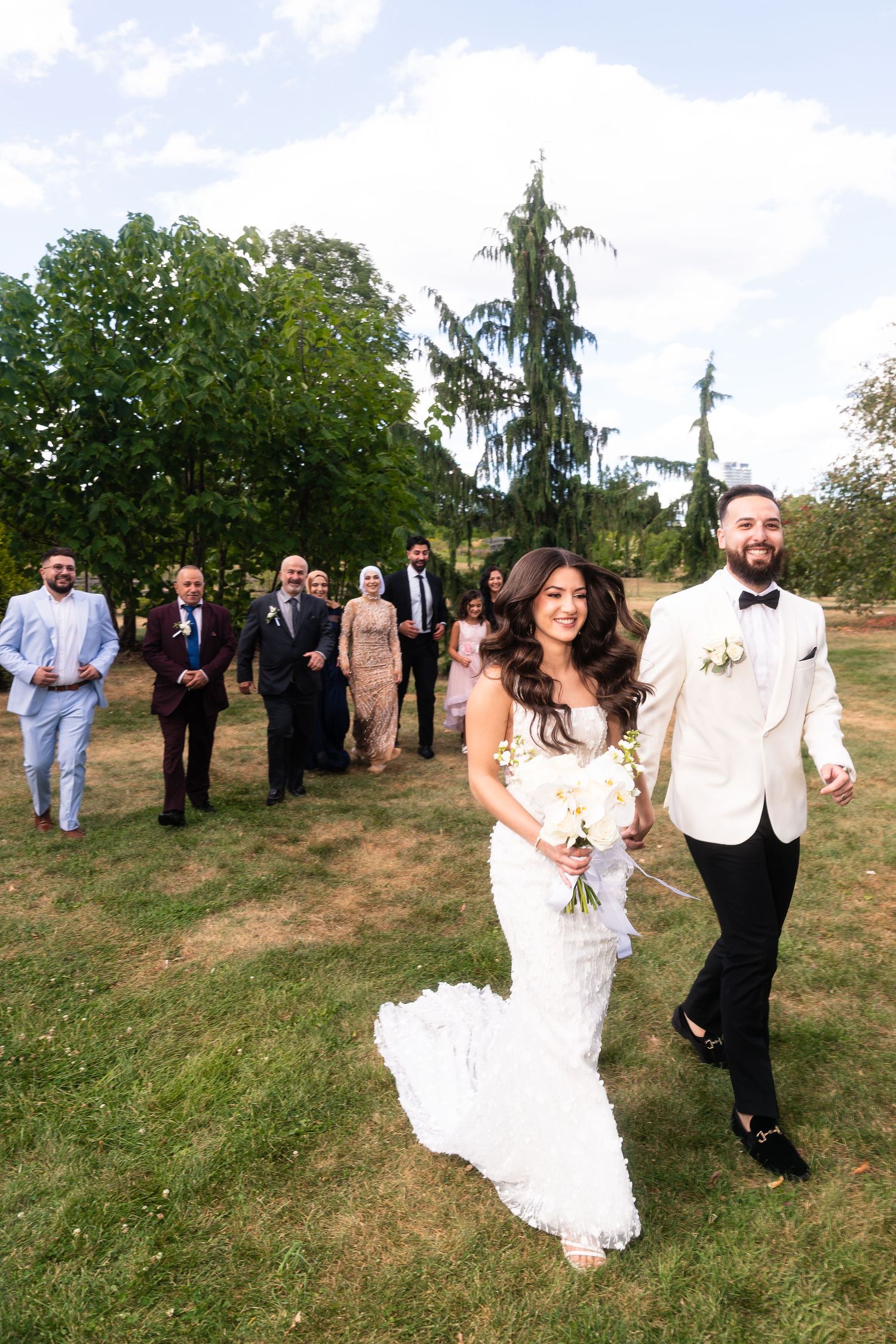 Newlyweds walking together across a grassy hill followed by their wedding party.