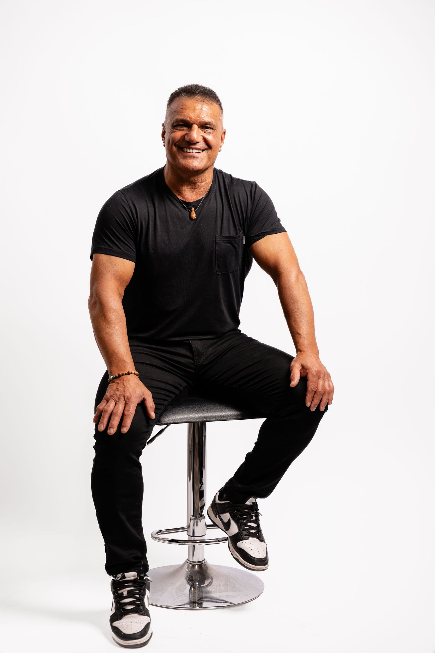 Athletic person in black t-shirt and pants sitting on modern chrome bar stool against white background.