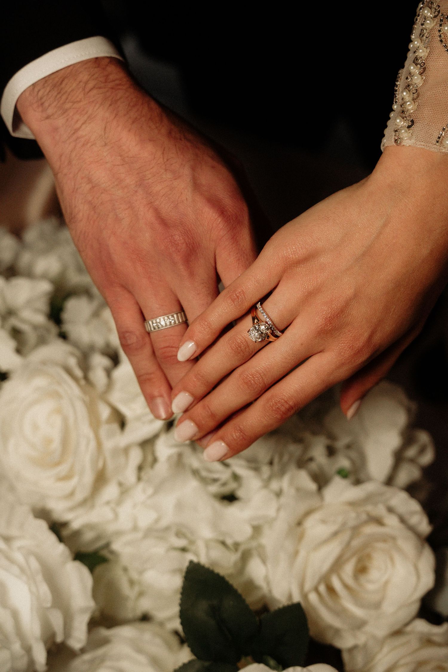 Close-up of hands with wedding rings resting on white roses.