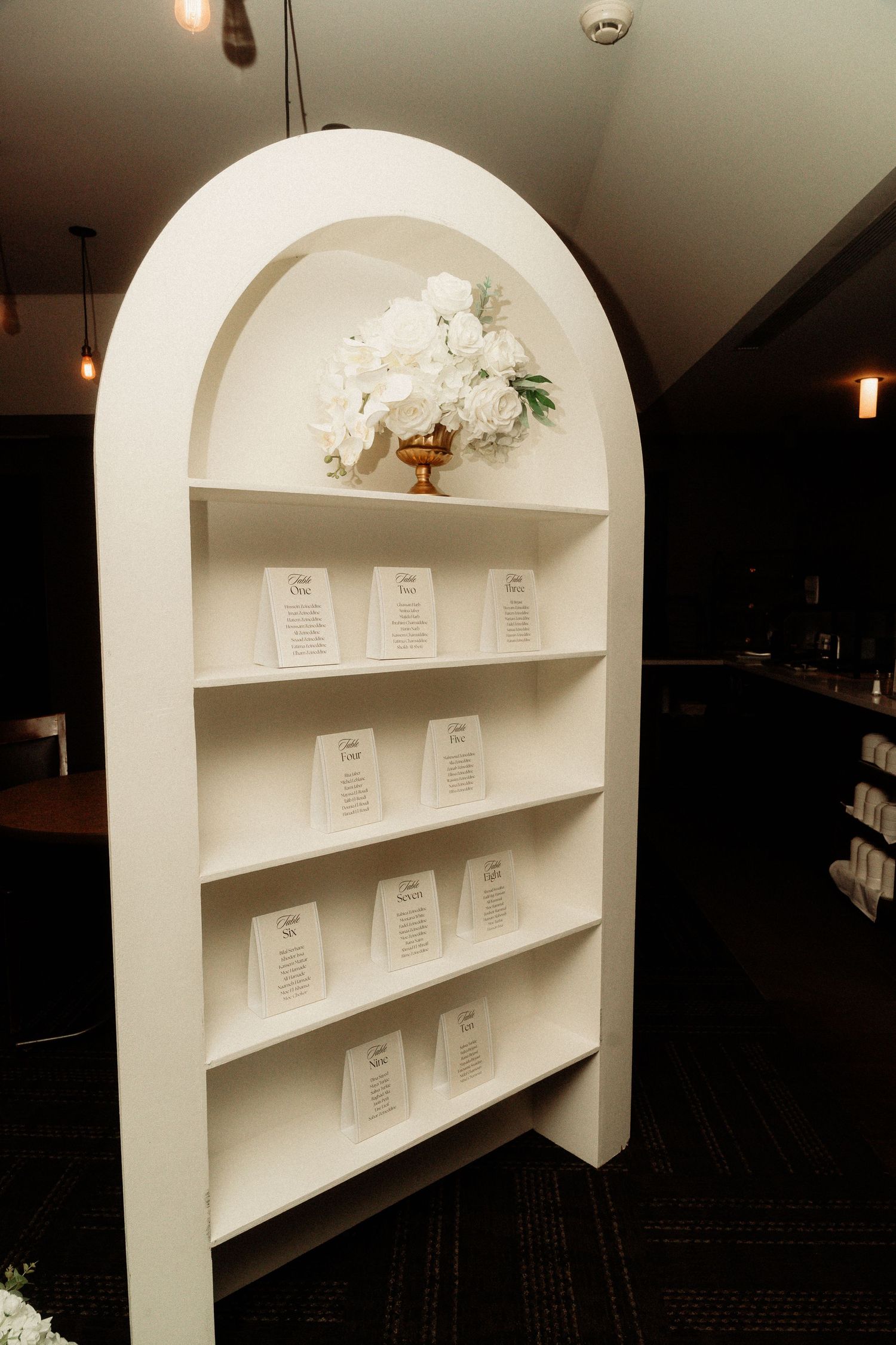Arched white shelving unit displaying wedding seating cards with floral decorations on top.