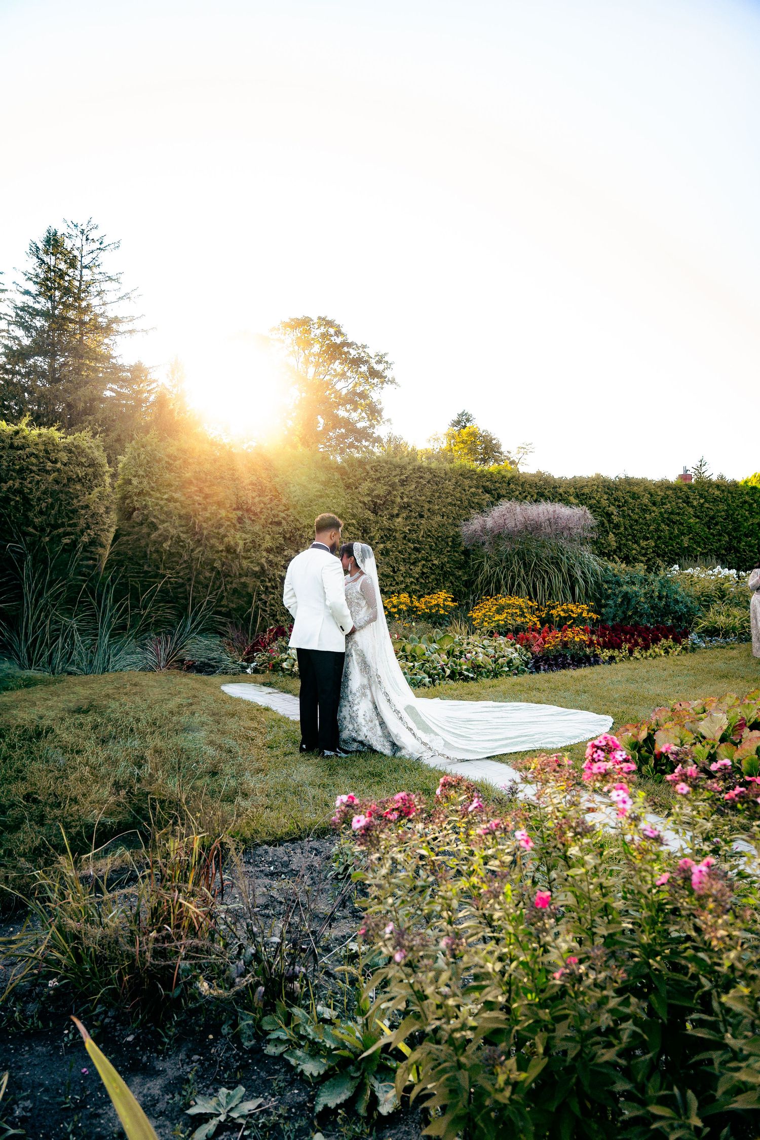 A couple poses in a garden at sunset with pink flowers and greenery creating a romantic outdoor setting.