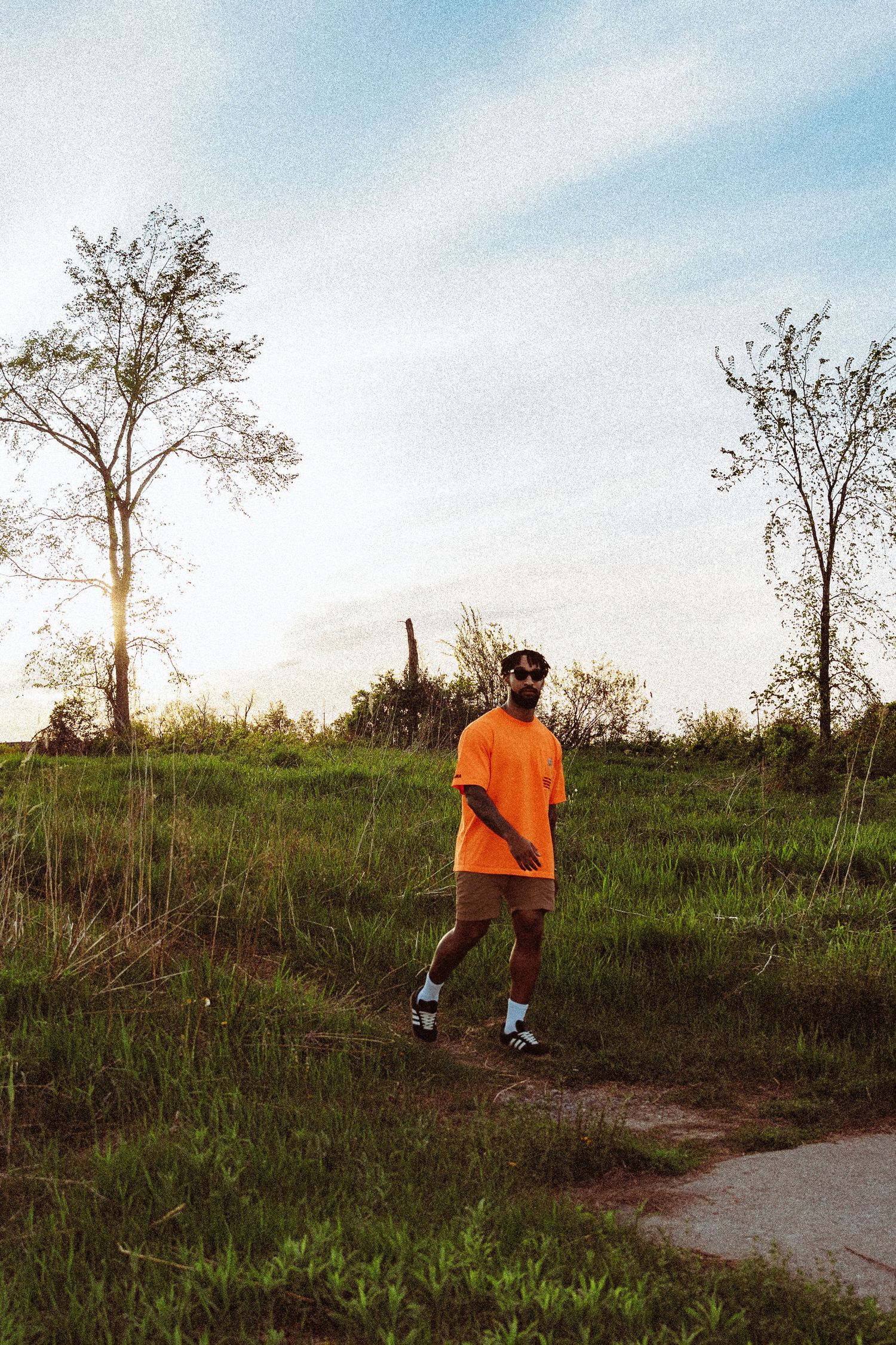 Person in bright orange shirt walking on dirt path through grassy field at sunset.
