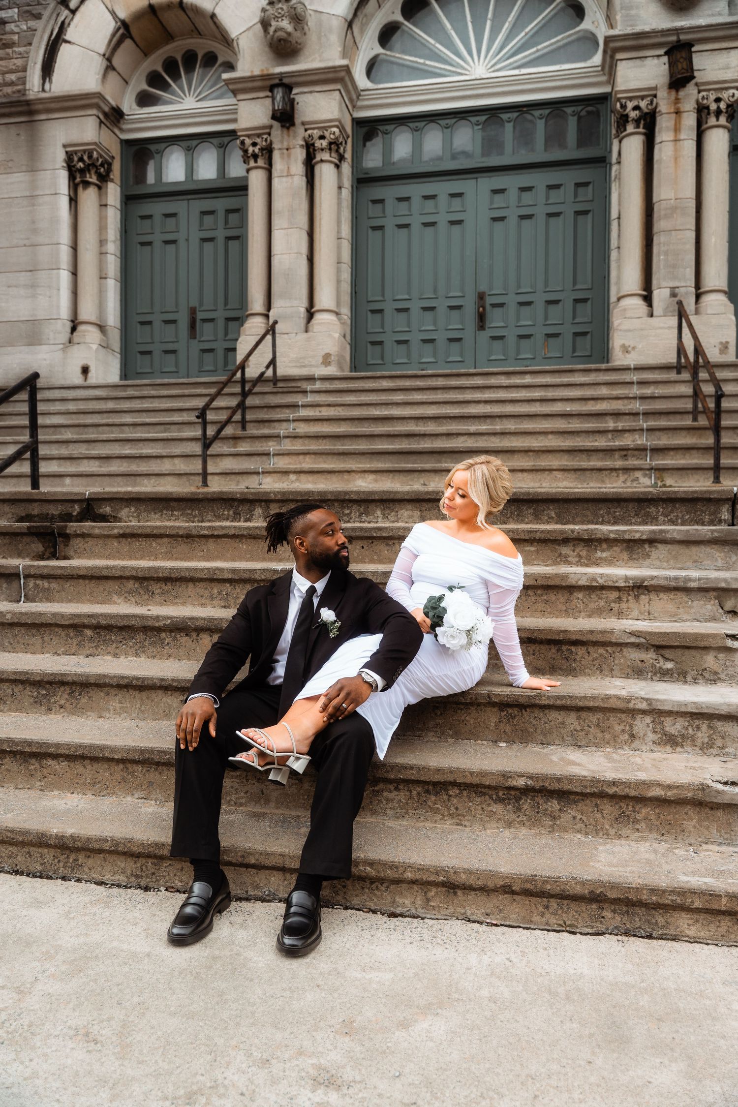 Wedding couple relaxes on grand stone steps of a historic building with green doors.