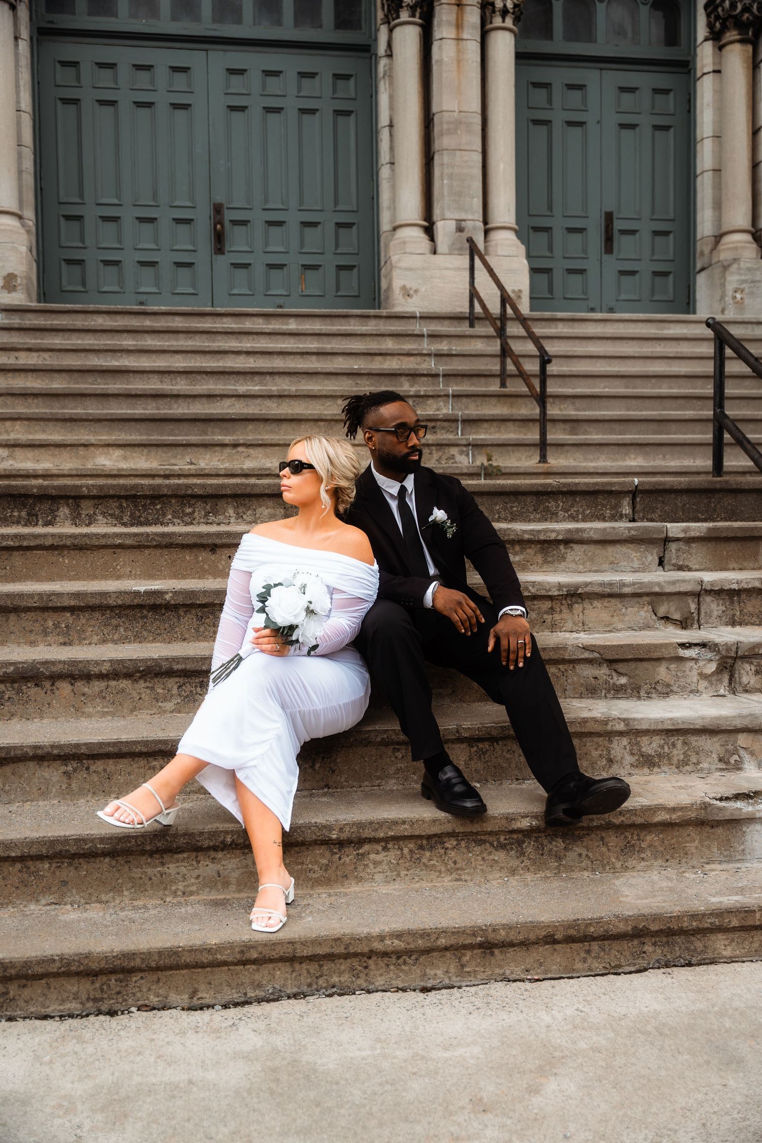 A bride in white dress and groom in black suit sit casually on stone church steps during their wedding photoshoot.