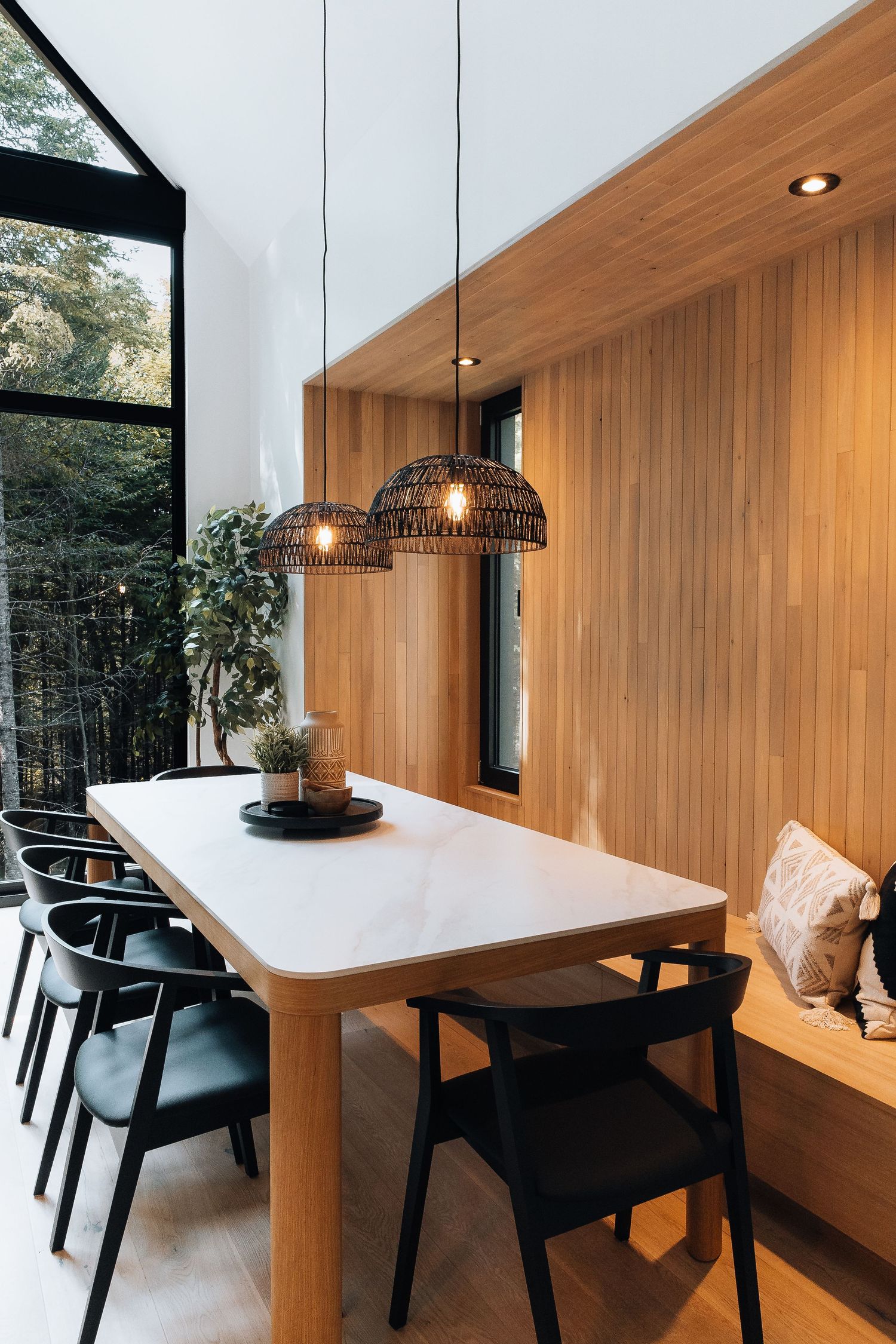 Dining area with white table, black chairs, and wood paneling features pendant lighting and forest views.