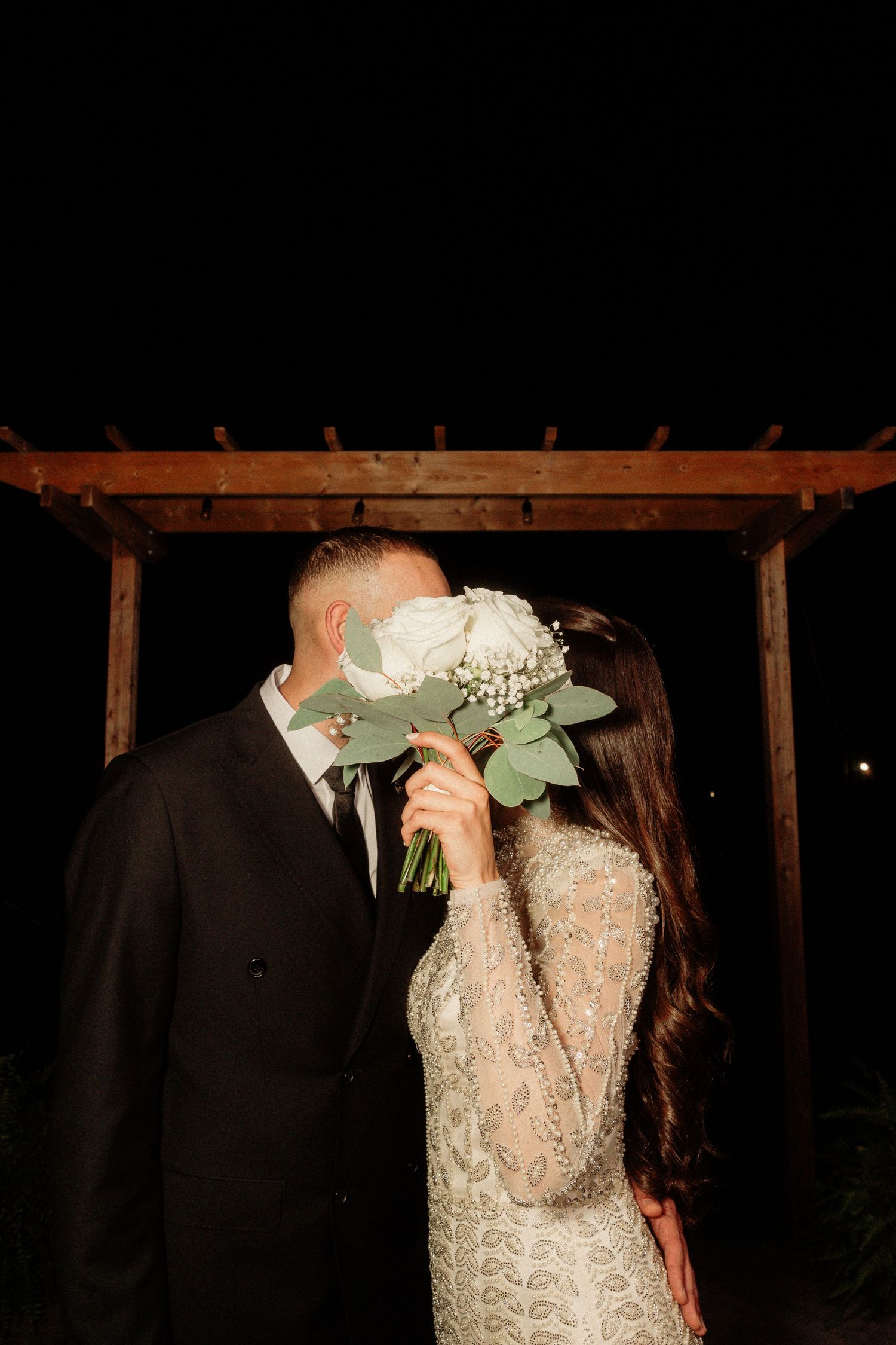 Romantic wedding couple sharing an intimate kiss under a rustic wooden pergola at night.