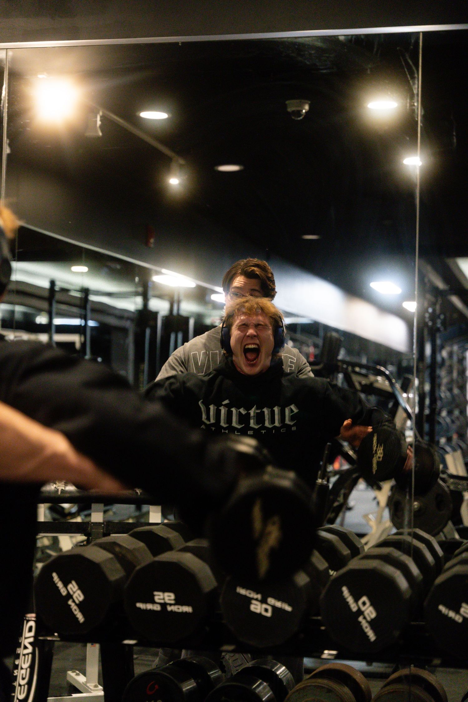 Person energetically posing over weight rack in gym wearing virtue branded shirt.
