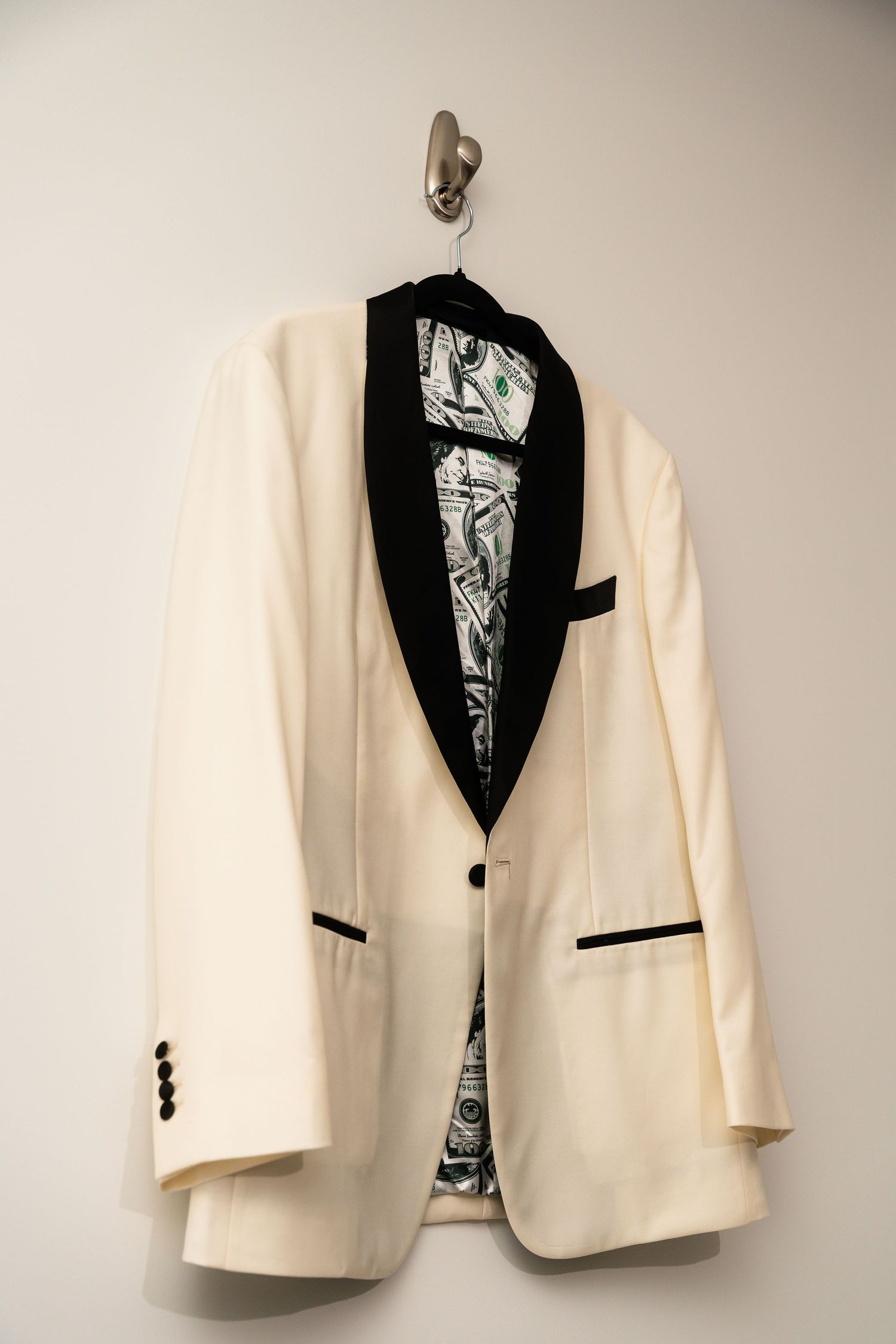A white formal dinner jacket with black lapels and trim hangs on a wall hook, displaying an elegant tuxedo design.