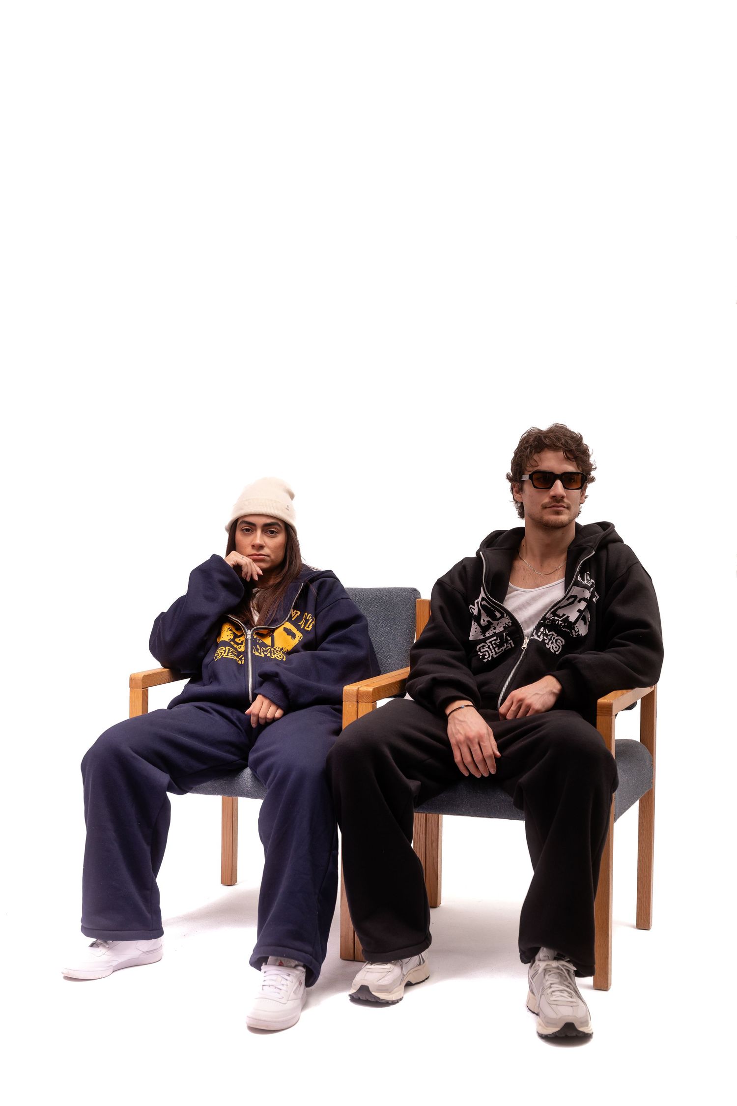 Two individuals seated on wooden chairs wearing casual streetwear.