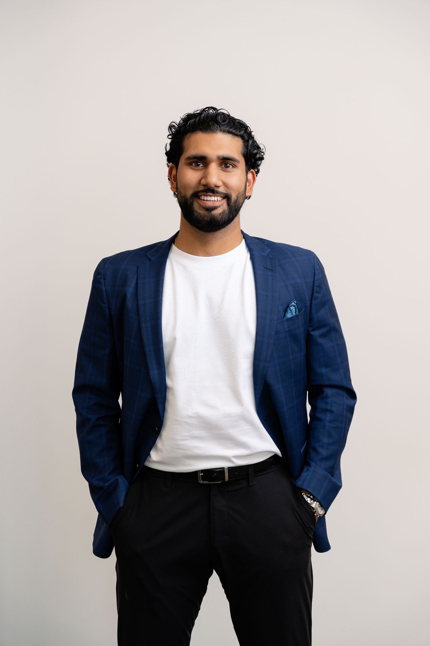 Professional in navy blazer and white t-shirt poses with hands in pockets against light background.