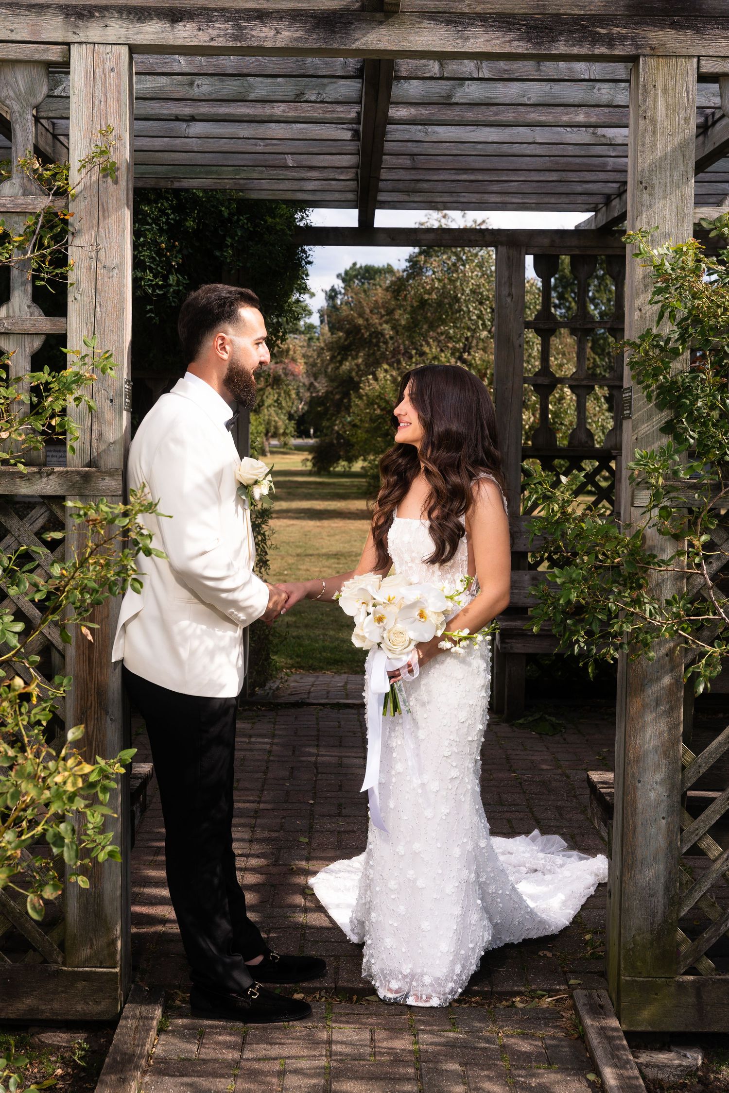 A couple shares a romantic moment under a wooden pergola in a garden setting during their wedding celebration.
