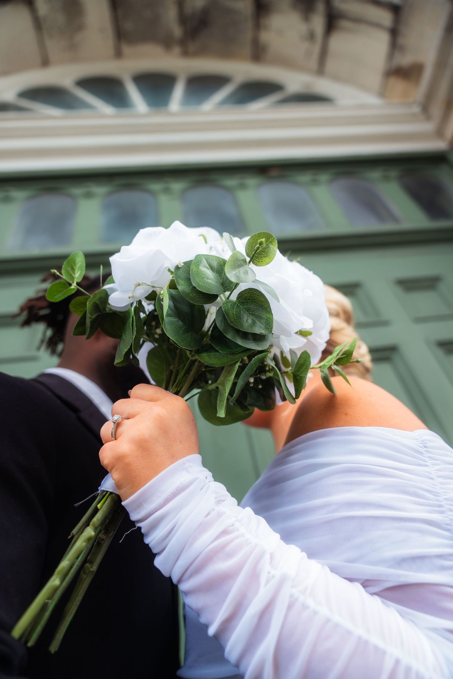 Close-up of hands holding a bridal bouquet with white flowers and eucalyptus leaves against a mint green door.
