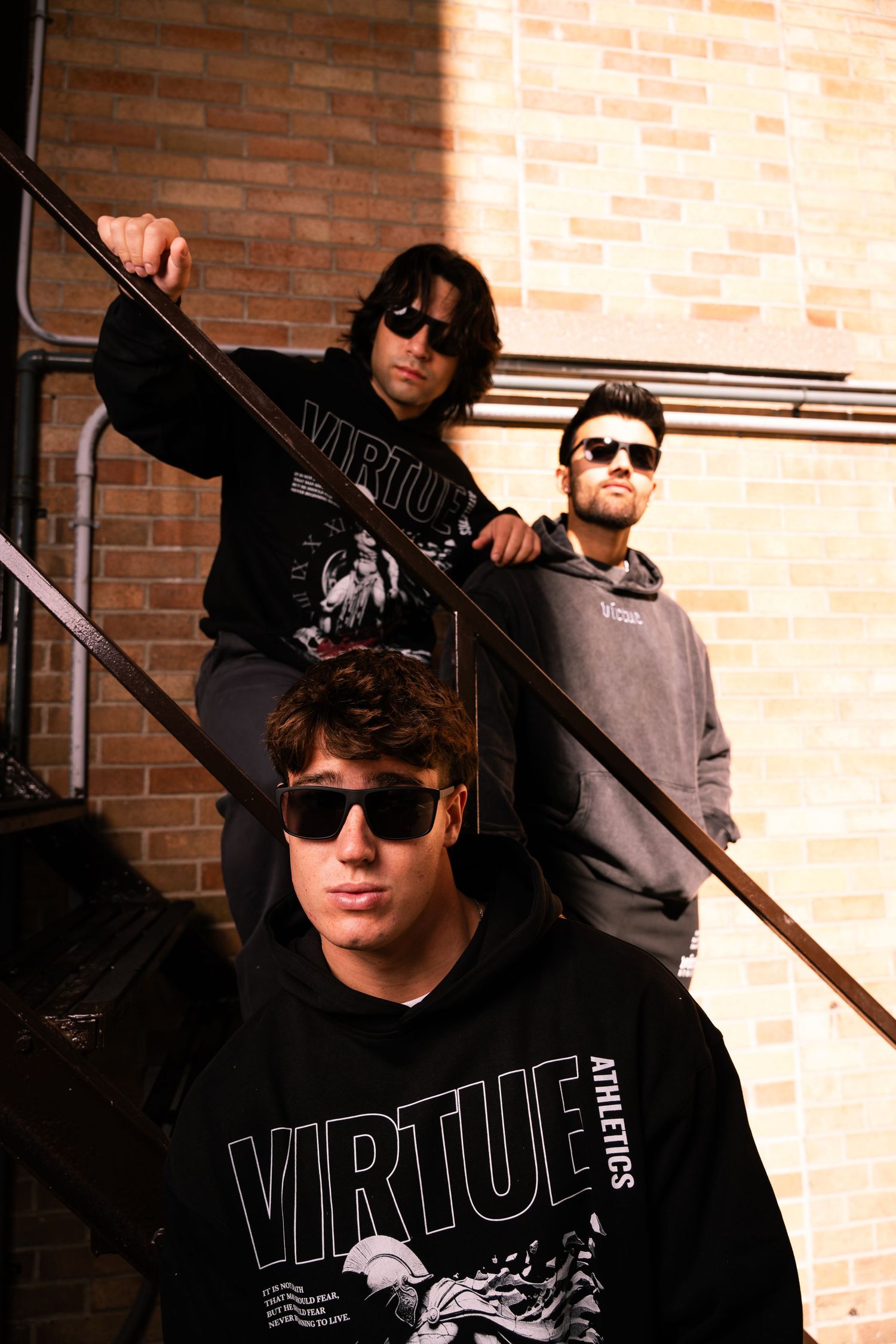 Group posed on brick building staircase wearing dark hoodies and sunglasses.