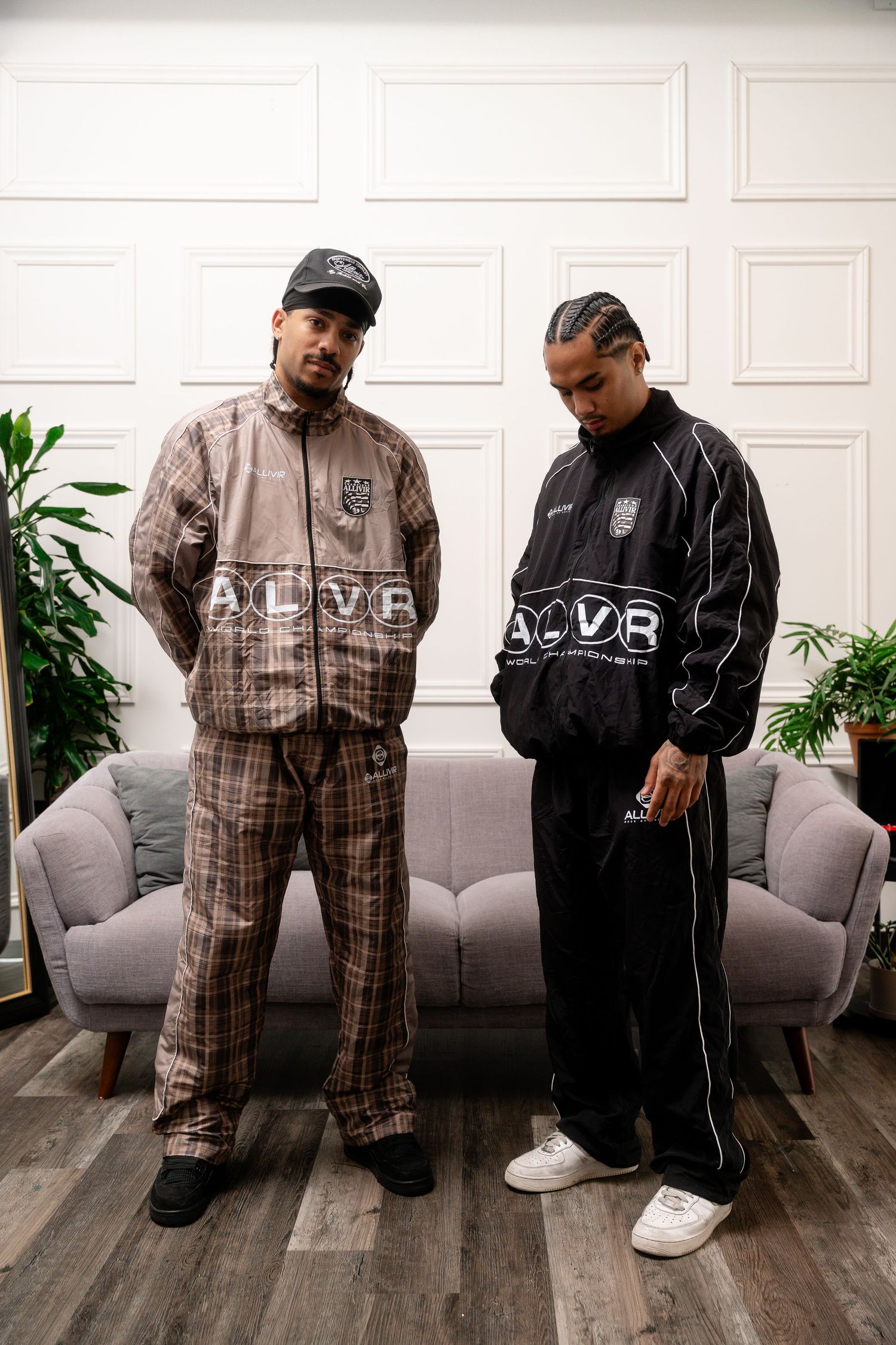 Two people wearing matching tracksuits posing in modern living room setting.
