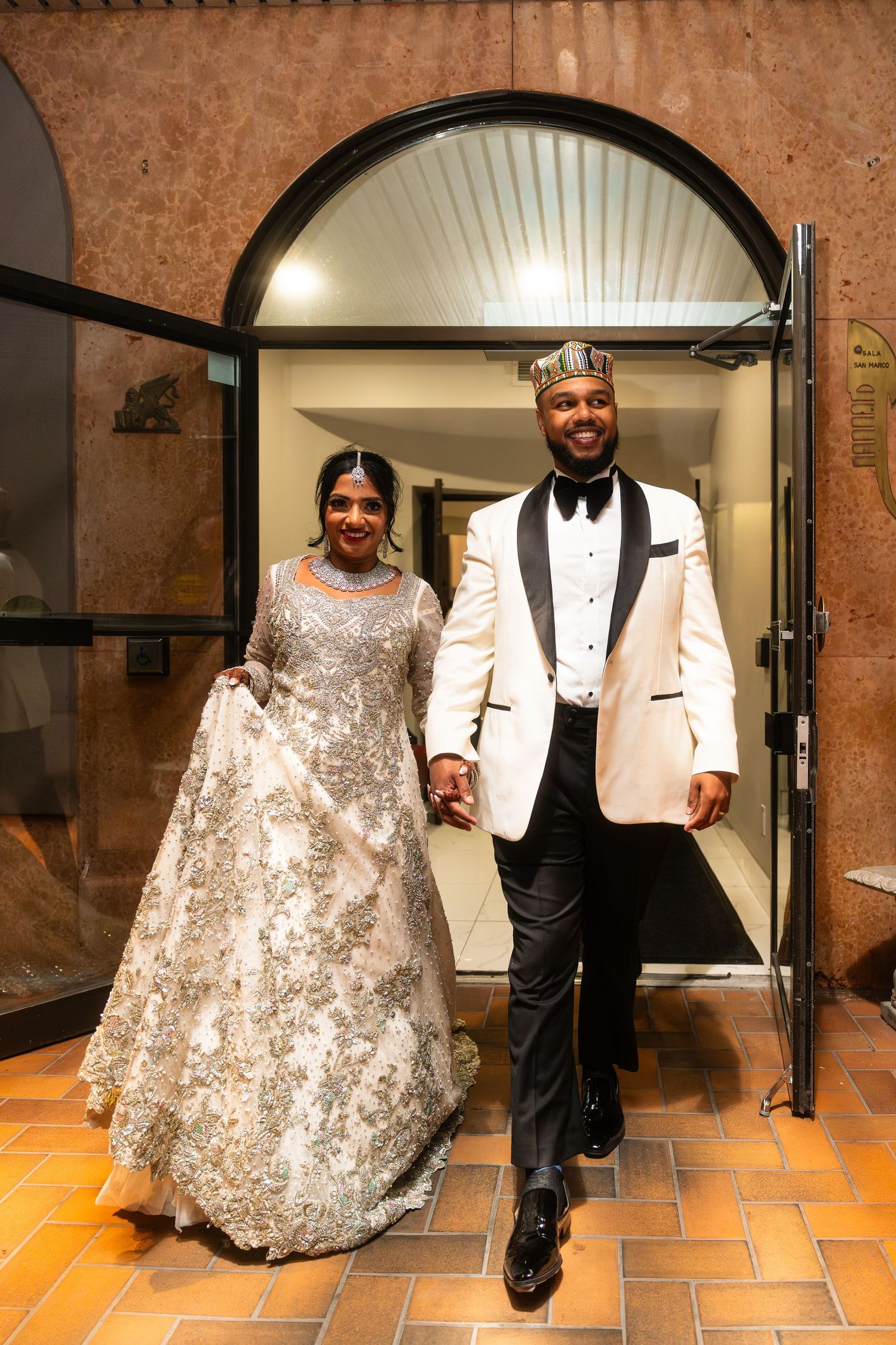 A couple in formal attire, with a white tuxedo jacket and sparkly silver gown, pose in an arched hallway.