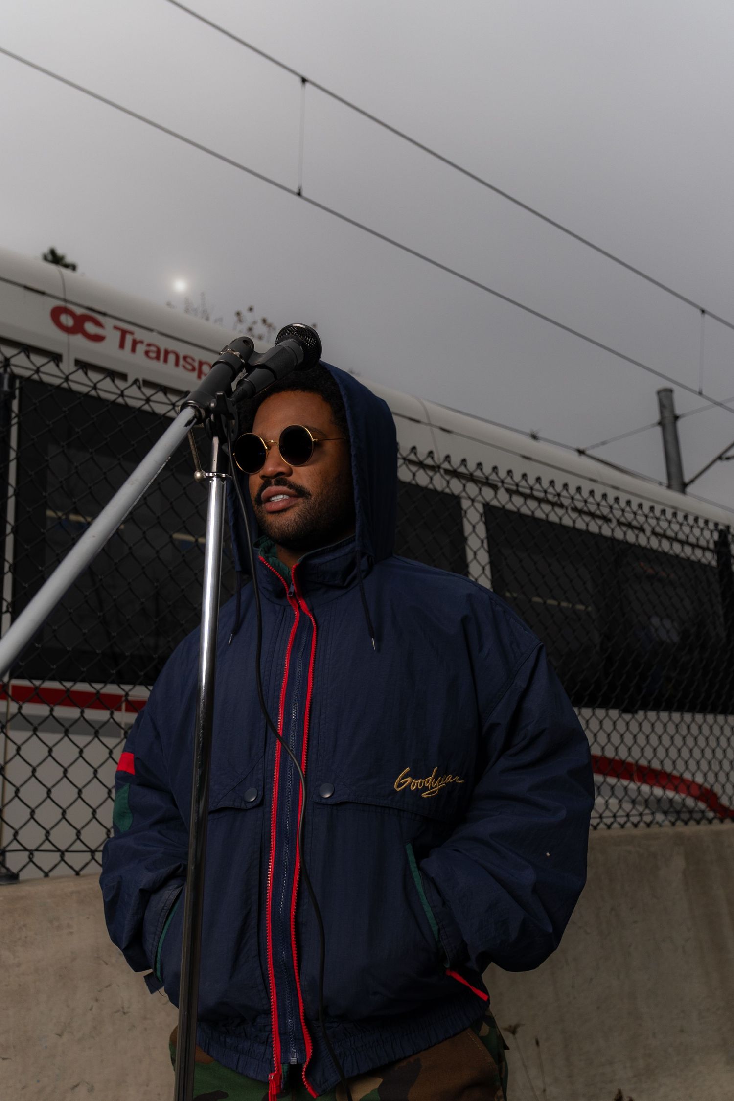Person in navy jacket and sunglasses posing against industrial fence backdrop.