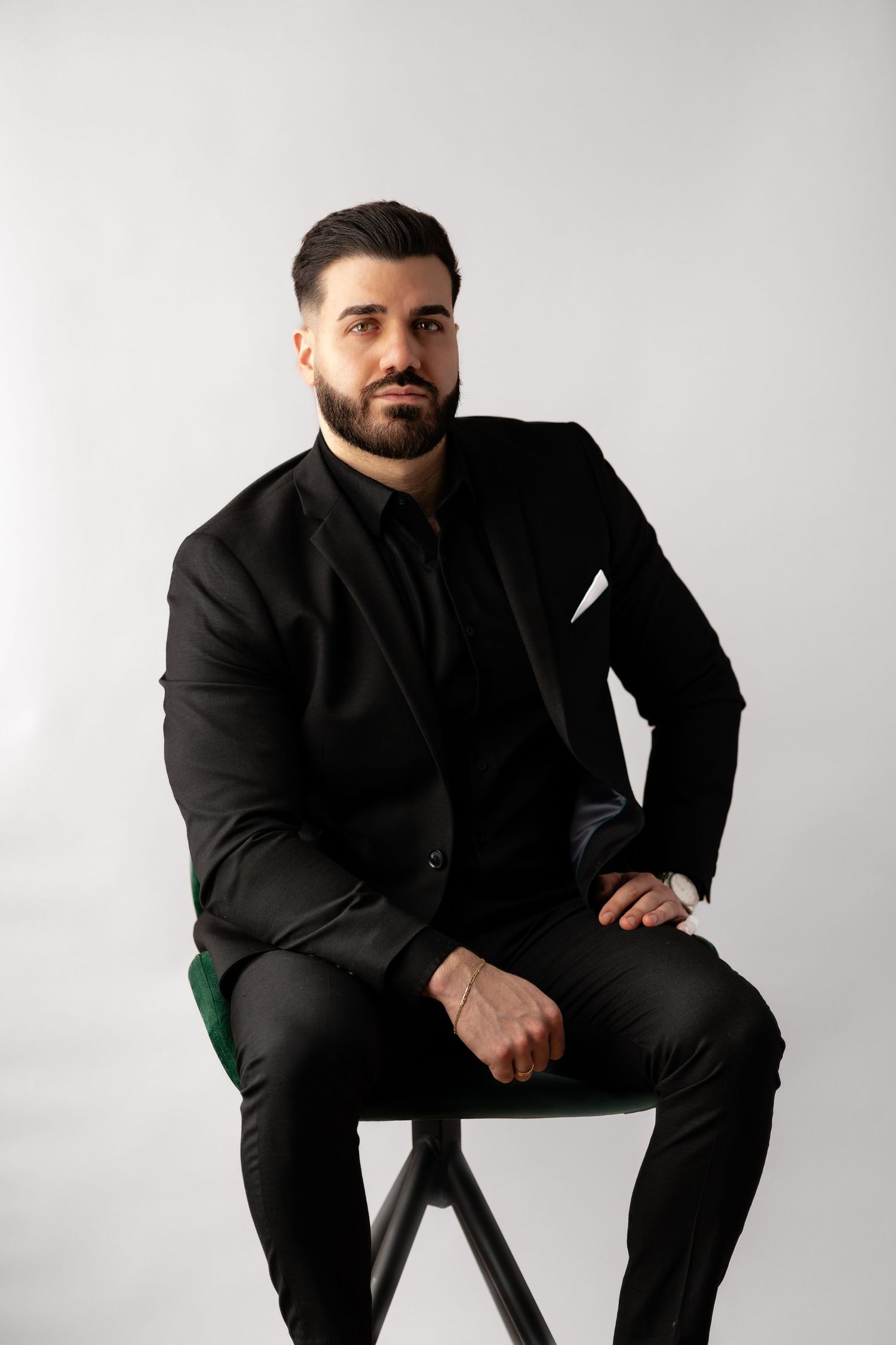 Man in black suit sitting on stool against light background in studio setting.