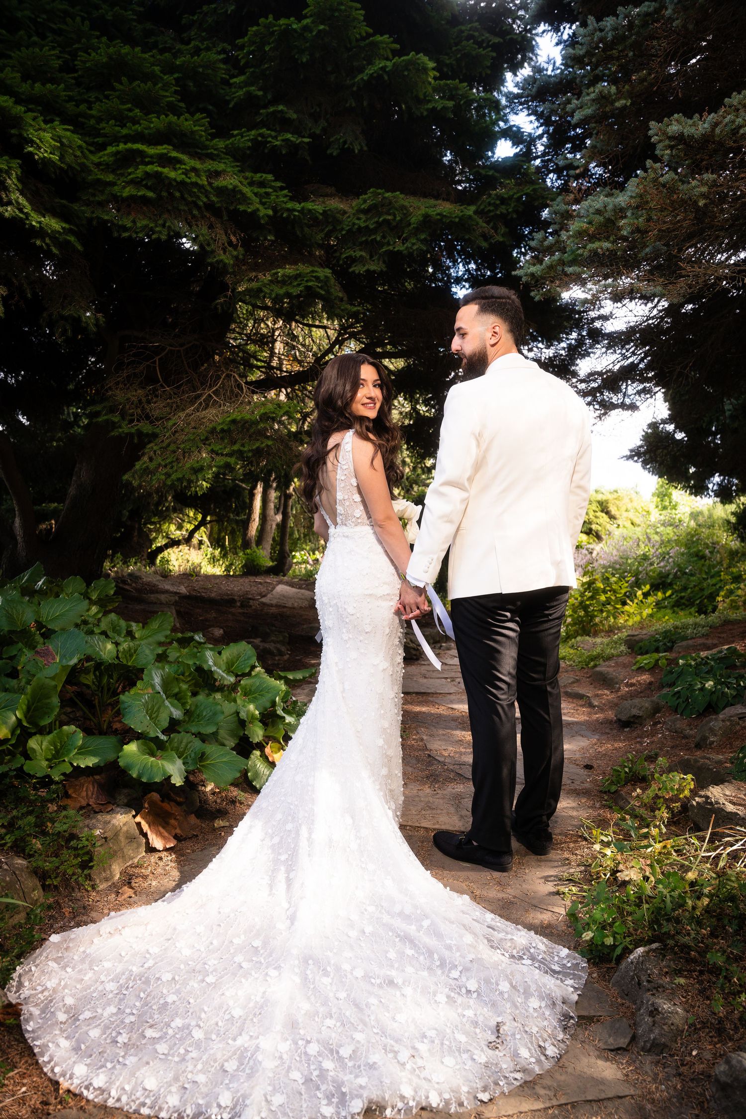 Wedding couple poses in elegant attire along wooded garden path.