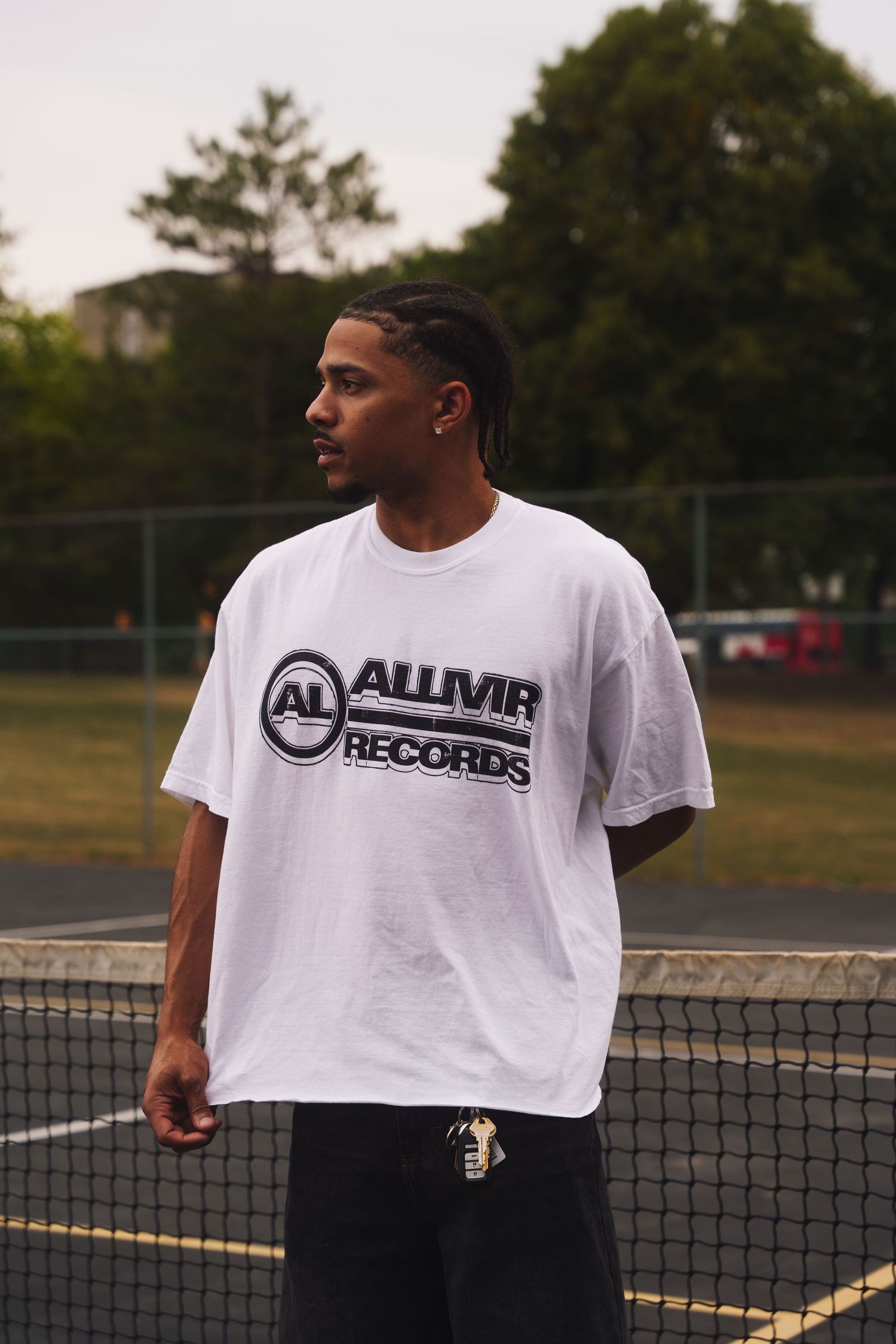 Person wearing ALLMVR Records logo t-shirt standing by tennis court net.