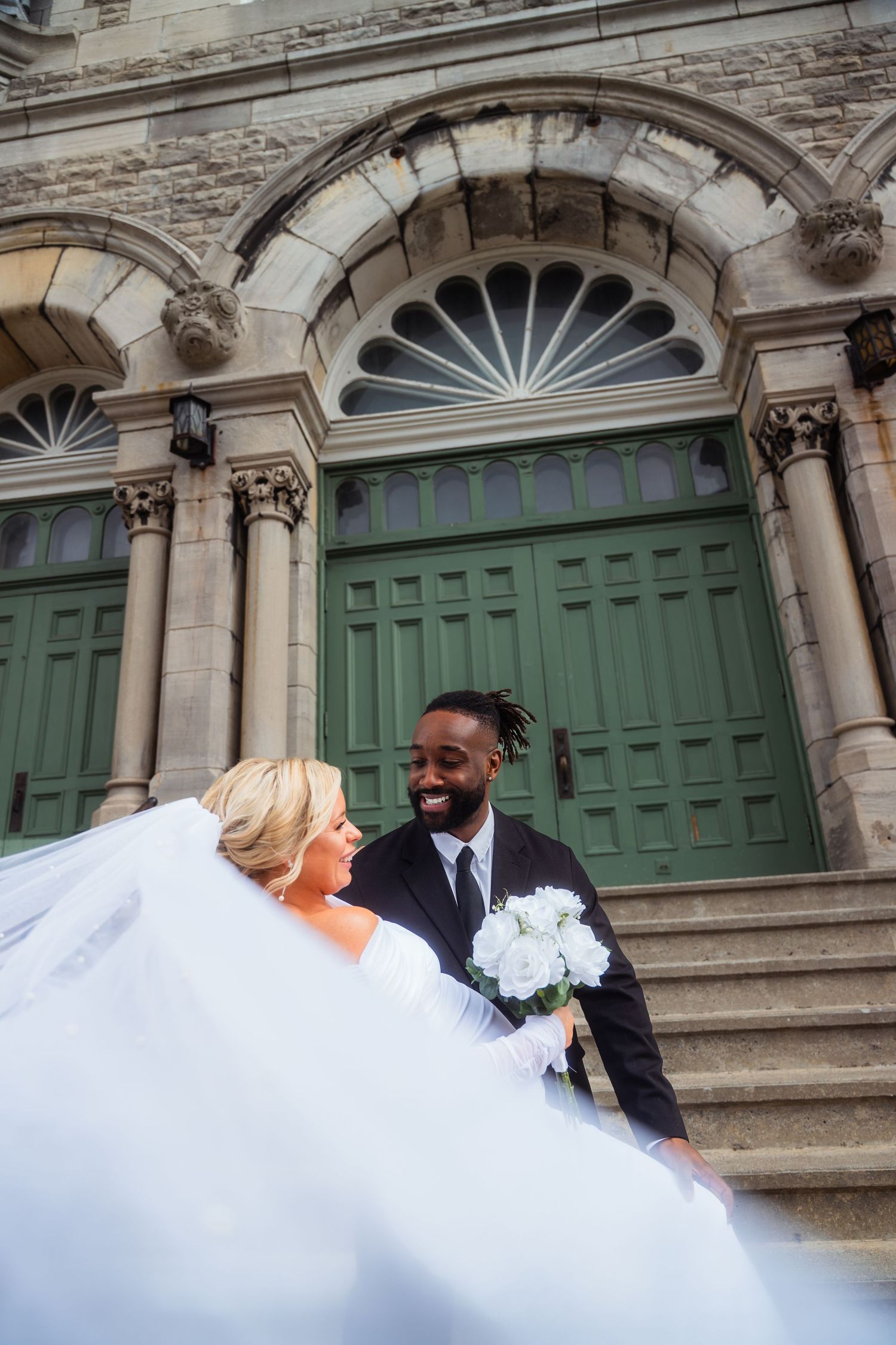 A wedding couple poses on the steps of a historic stone building with green doors and ornate architectural details.