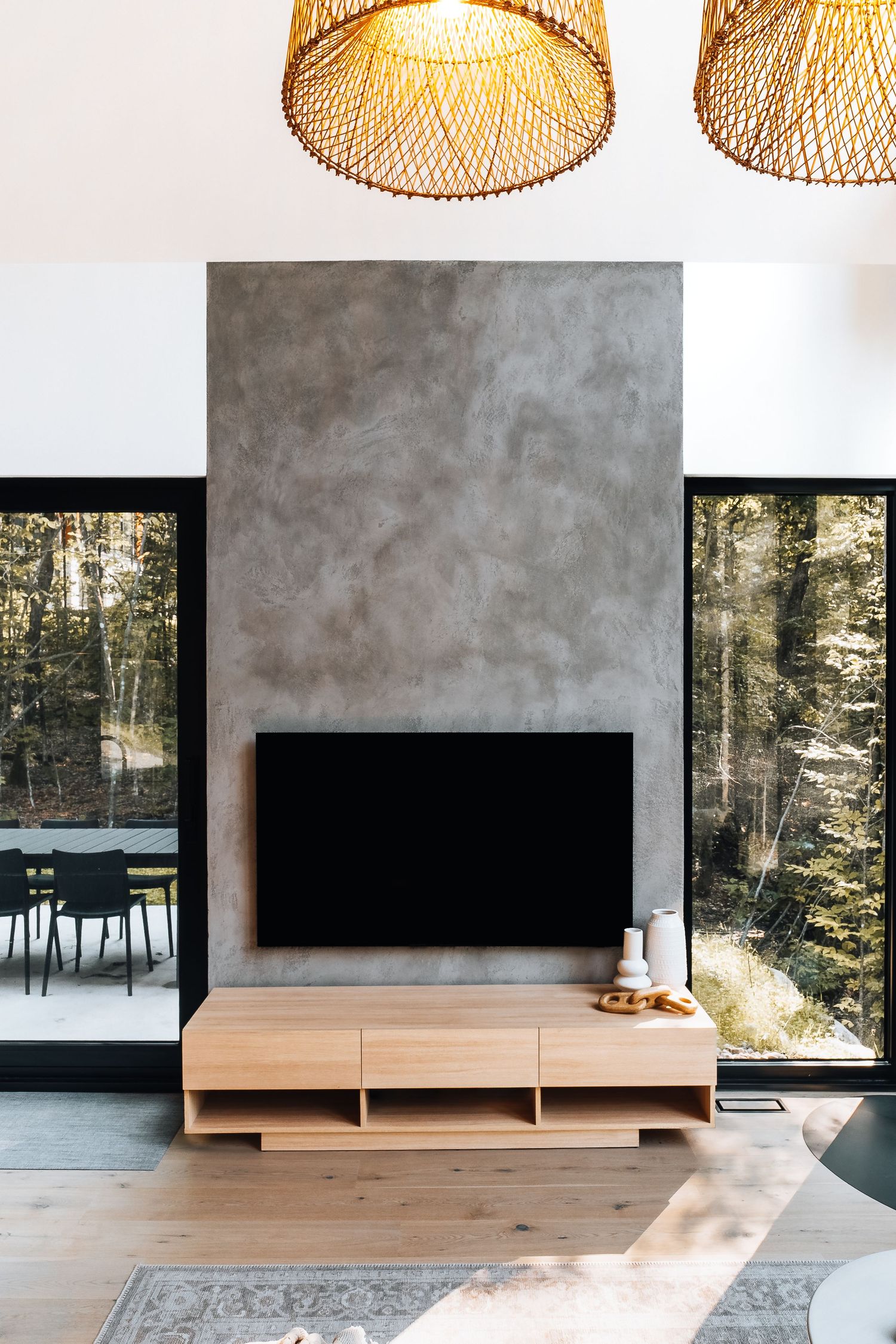 Minimalist living room with concrete fireplace, wooden media console, and large windows overlooking forest.