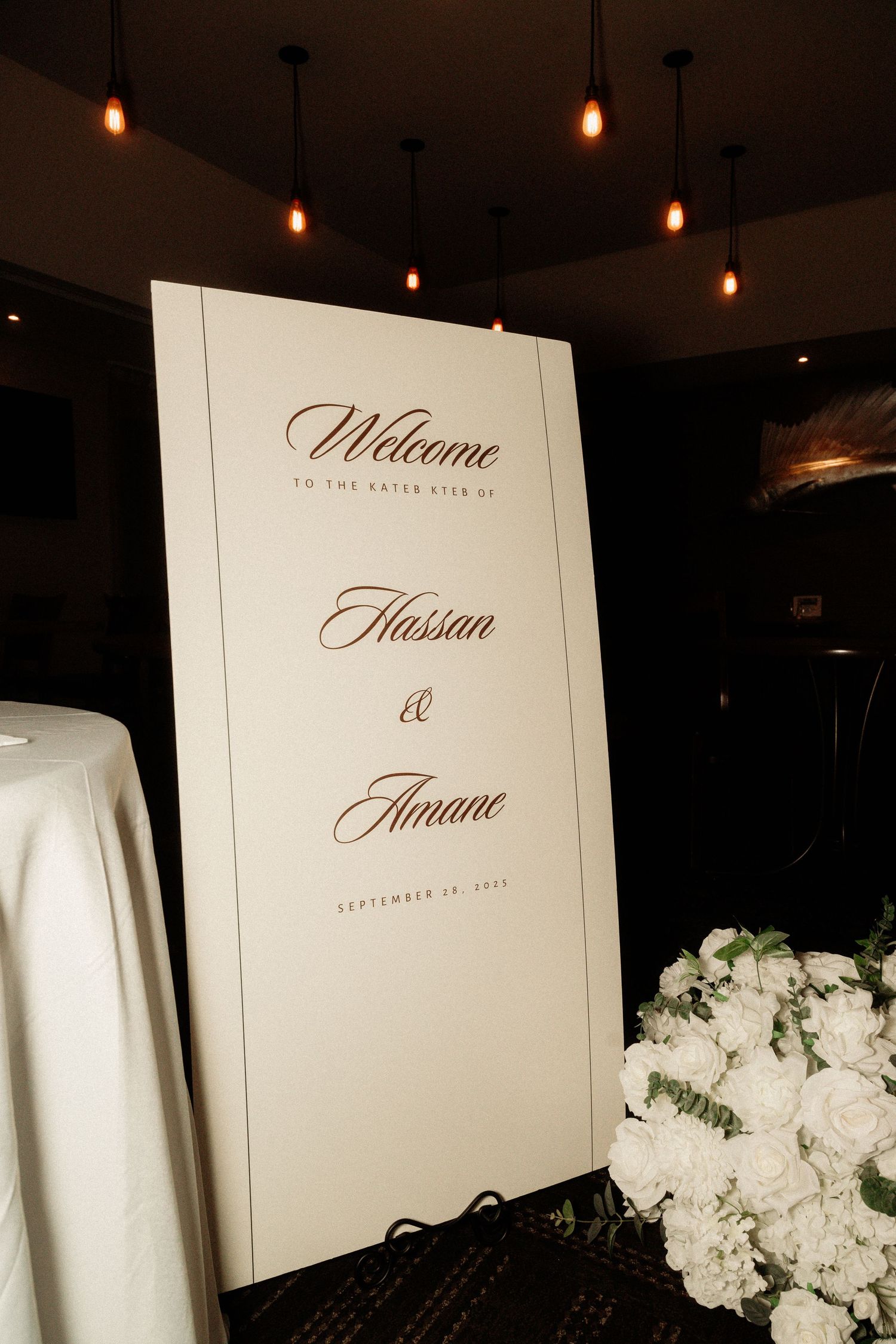 Elegant wedding welcome sign with calligraphy text and white floral decorations under pendant lights.