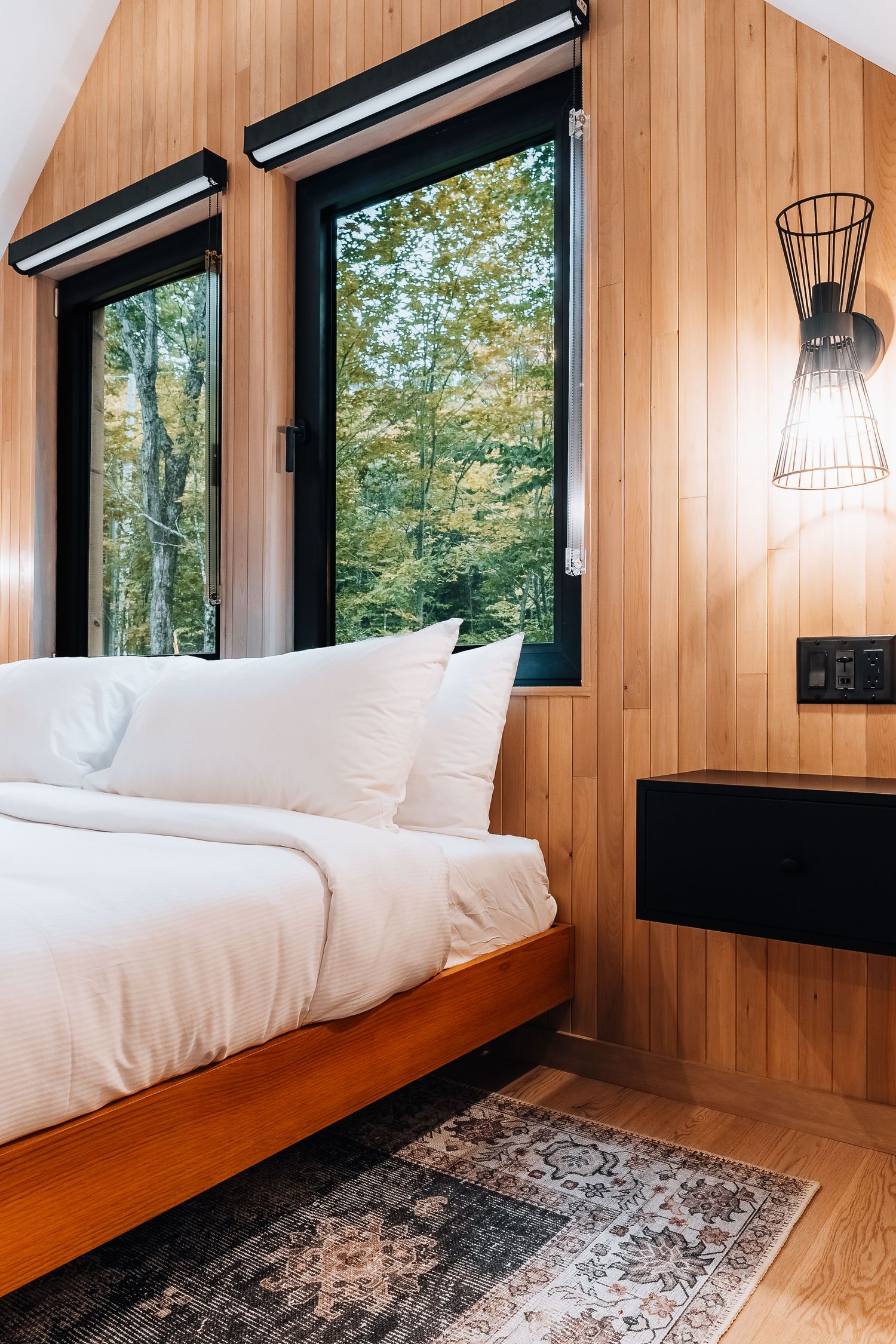 Cozy bedroom with wood-paneled walls, large windows, and modern sconce lighting.