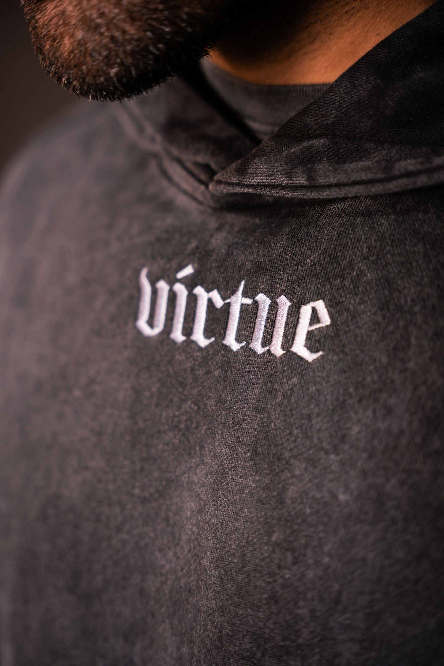 Close-up view of black hoodie with 'virtue' text embroidered in white gothic lettering.