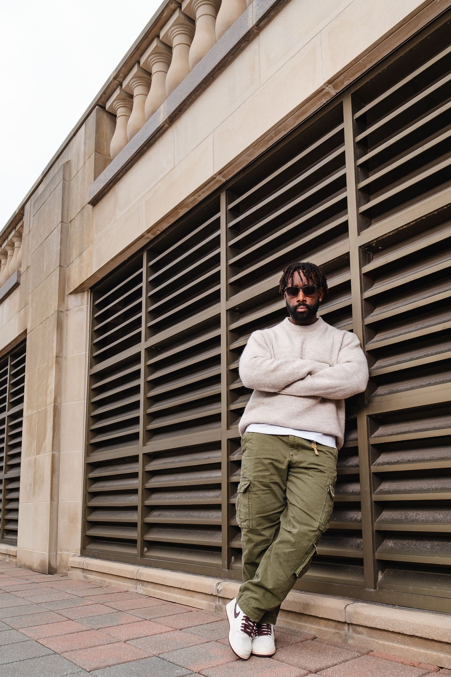 Person in white sweater and olive pants sitting against industrial building wall.