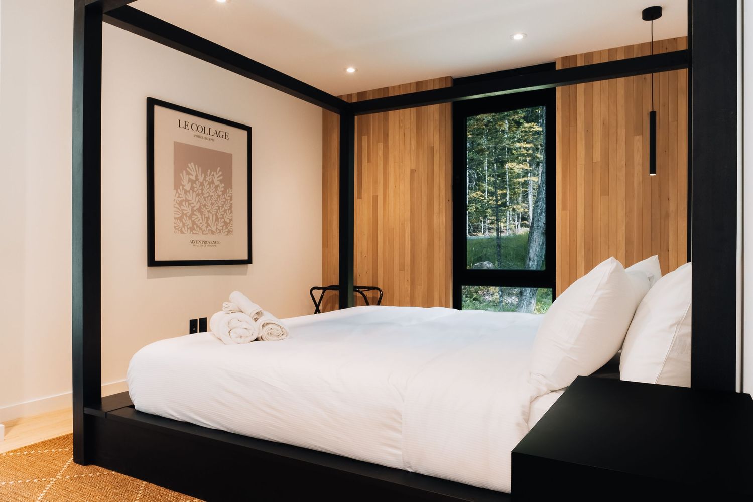 Minimalist bedroom with white bedding and wooden accent wall features large windows overlooking forest.