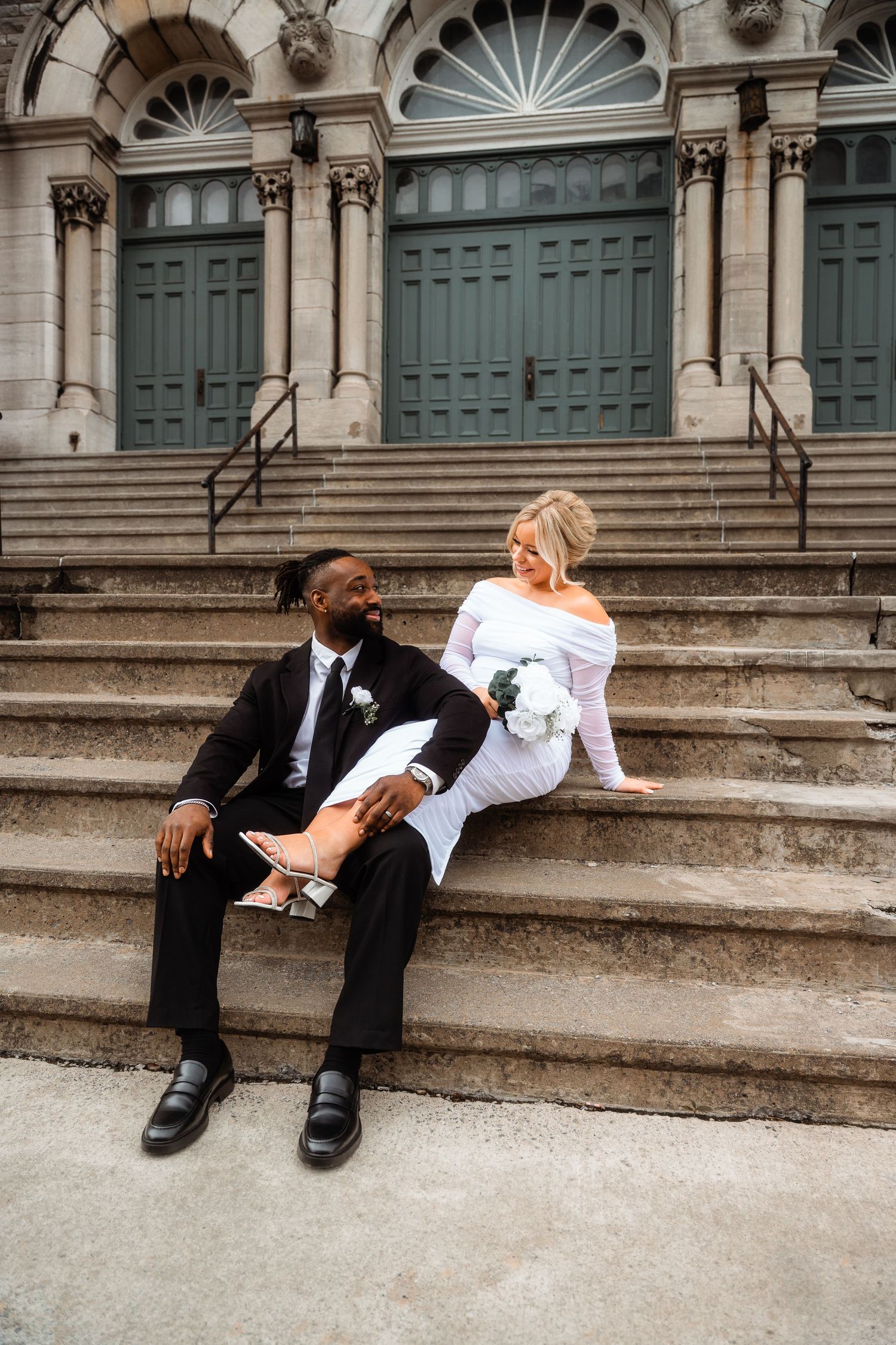 A couple relaxes casually on grand stone church steps with elegant green doors in the background.