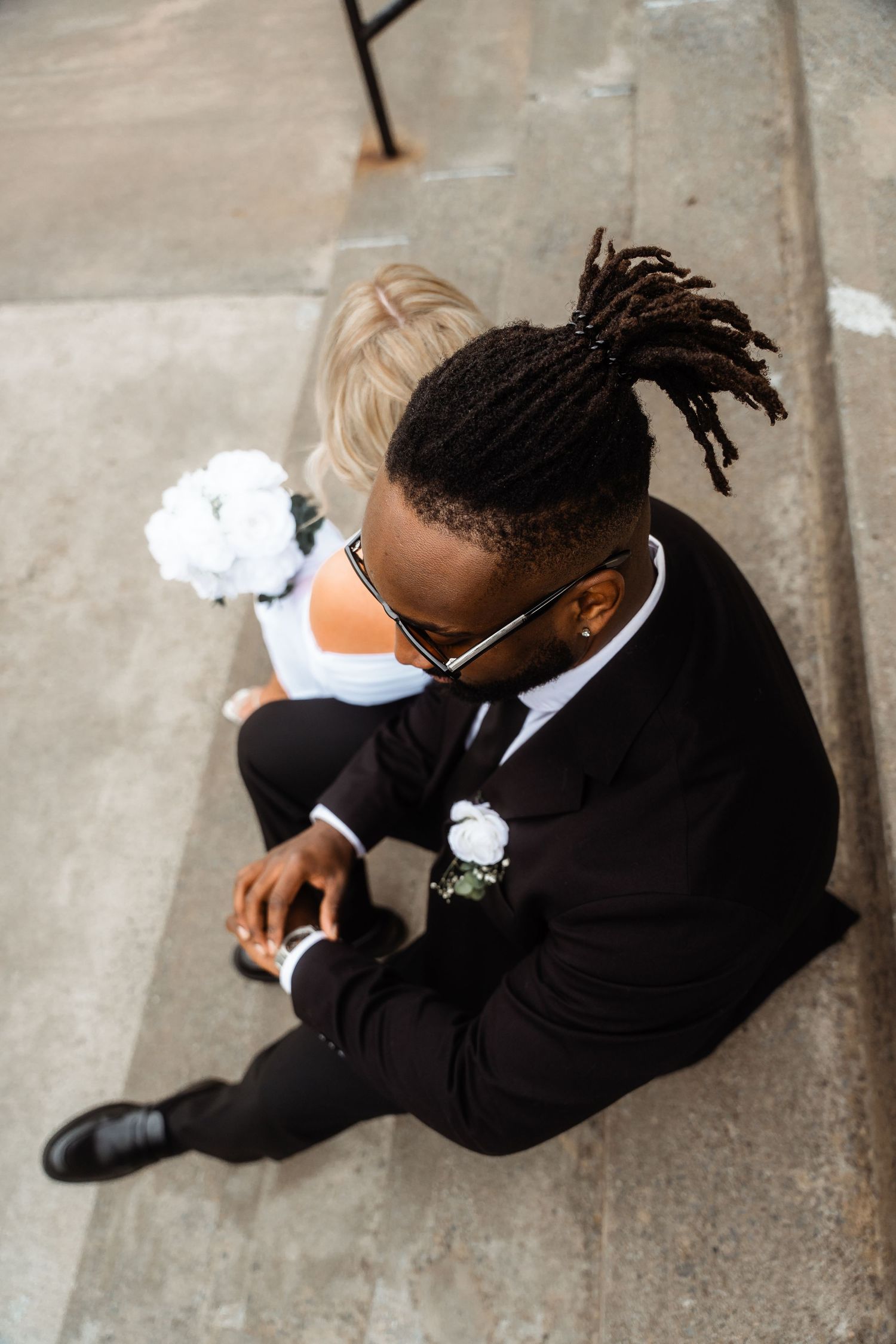 An overhead view of a groom in classic black tuxedo sitting on stone church steps.