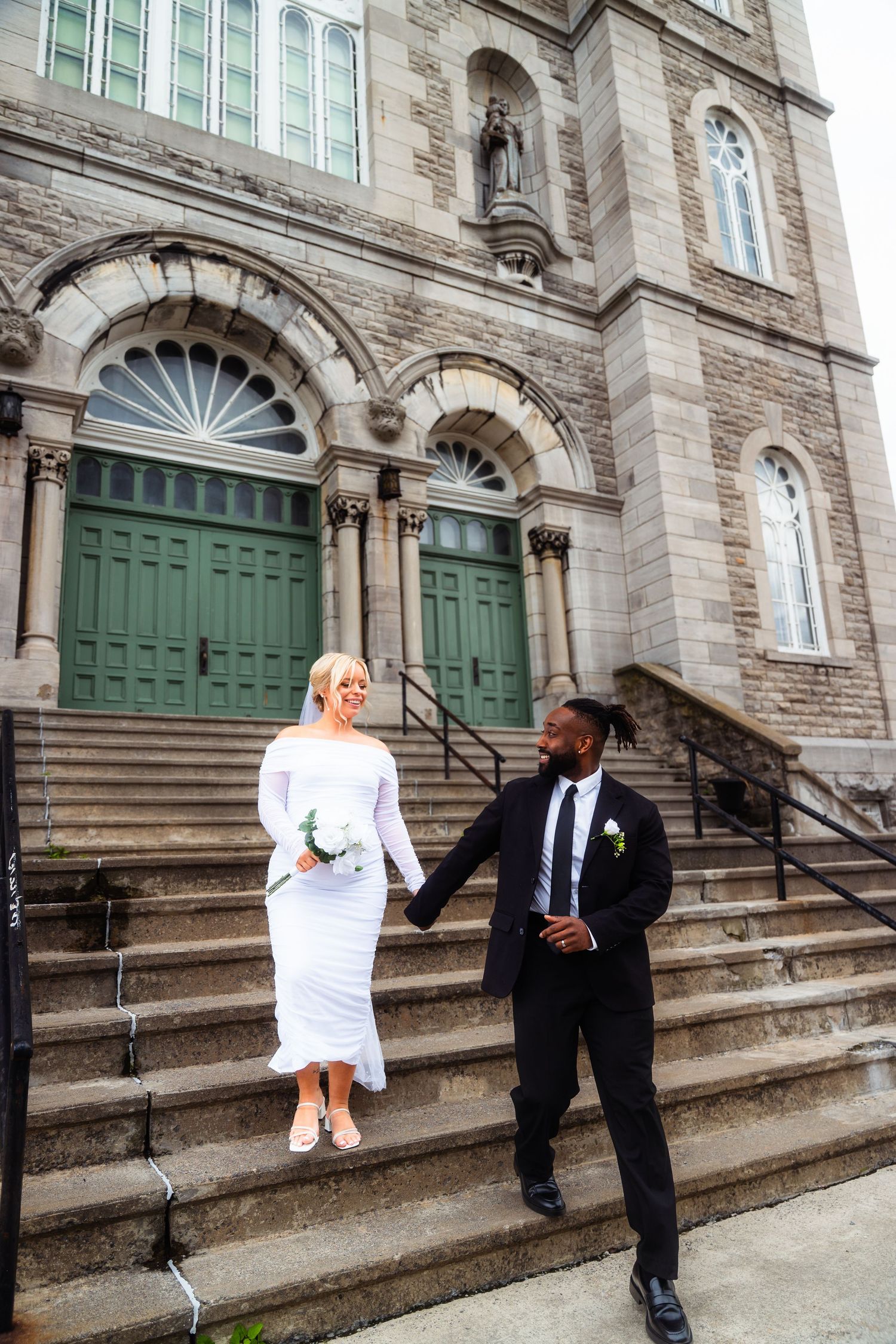 A couple stands on stone church steps with elegant green doors and arched architecture in the background.
