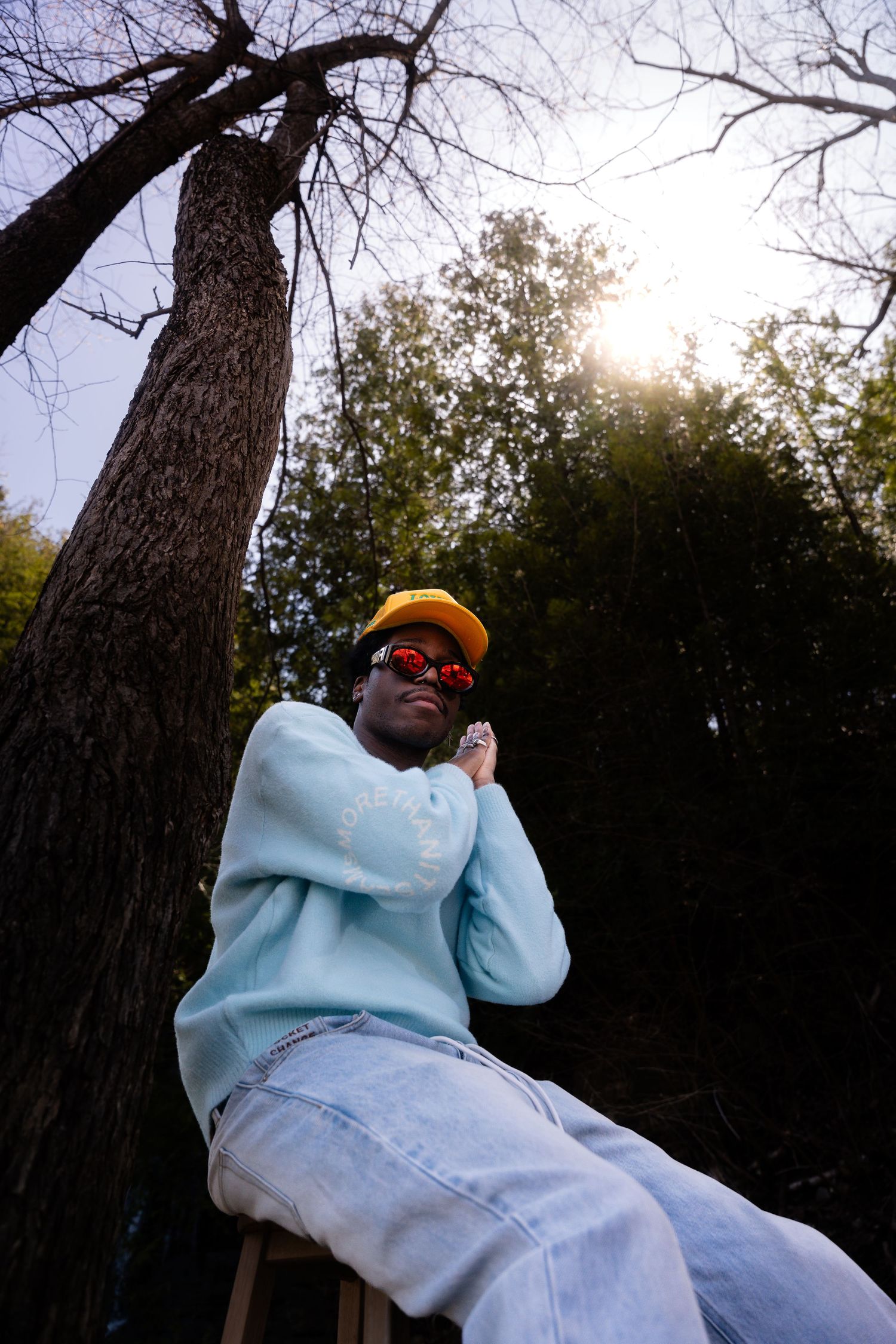 Person in light blue sweater and yellow cap sitting against tall tree trunk at sunset.