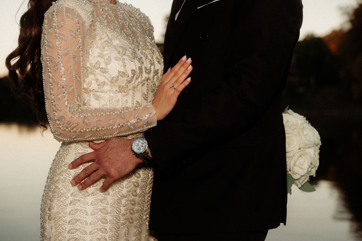 Couple in formal wedding attire embracing during sunset.