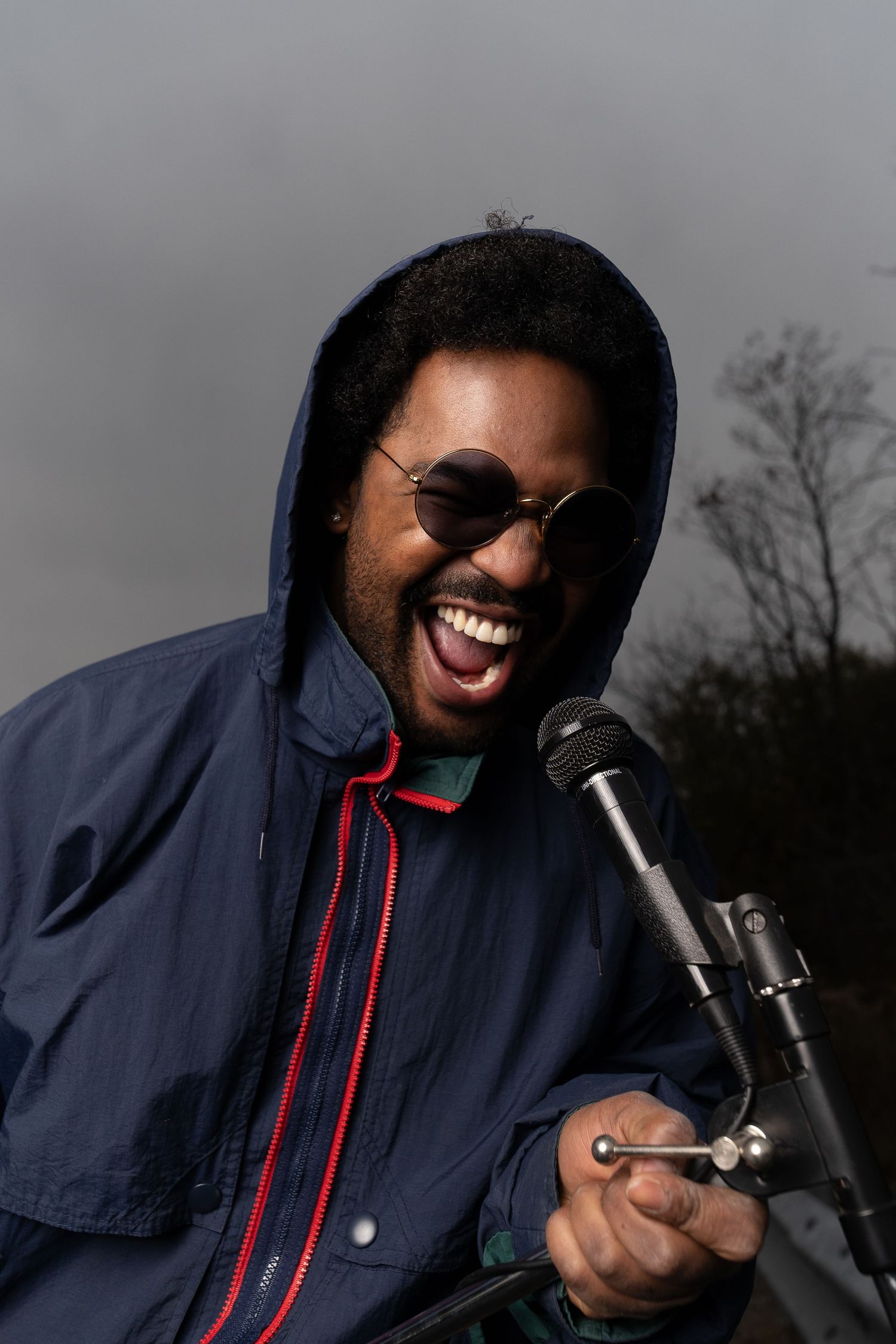 Person in navy hoodie laughing while holding microphone outdoors.