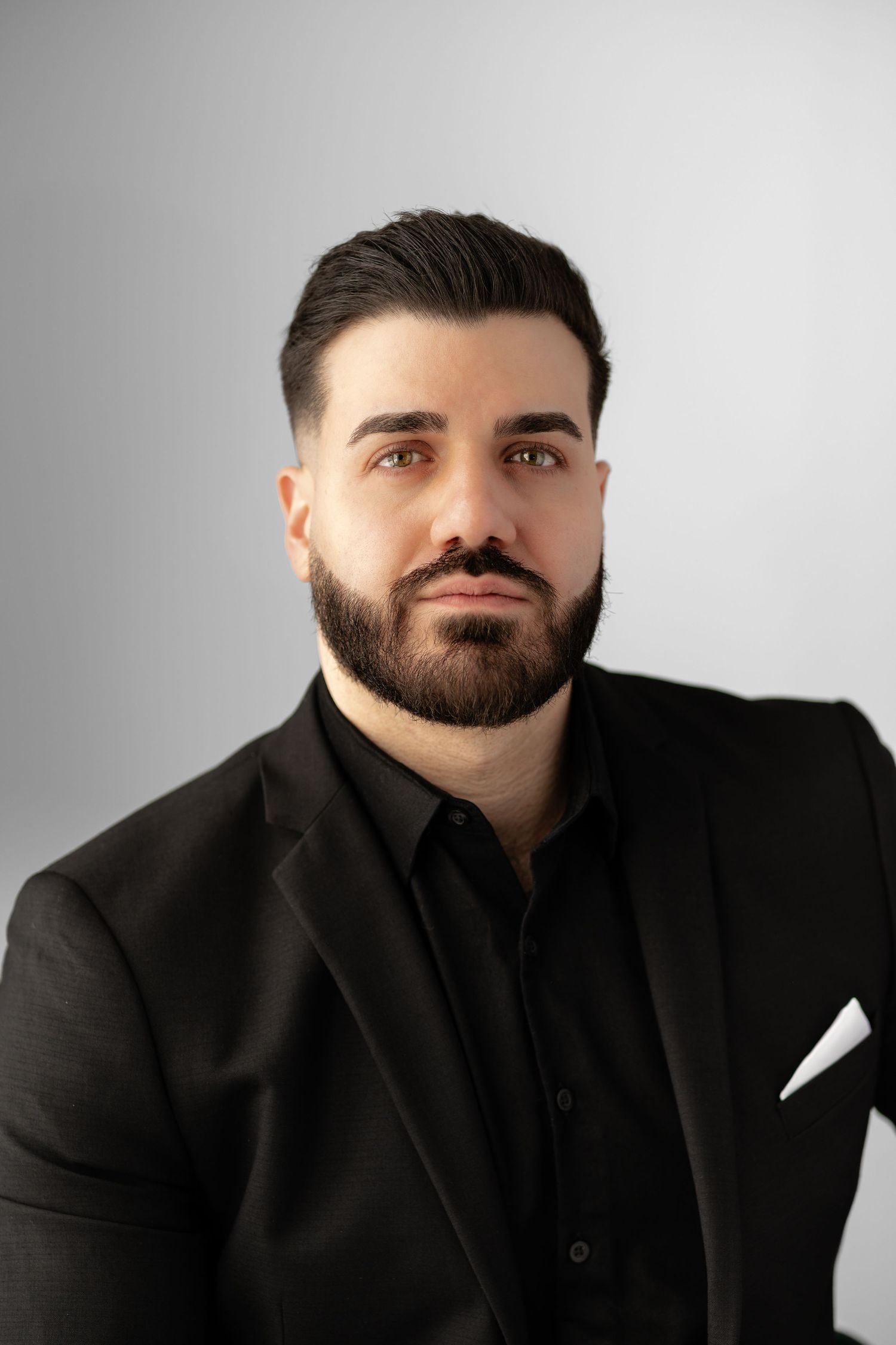 Professional headshot of a bearded man in a black suit and shirt against a light gray background.
