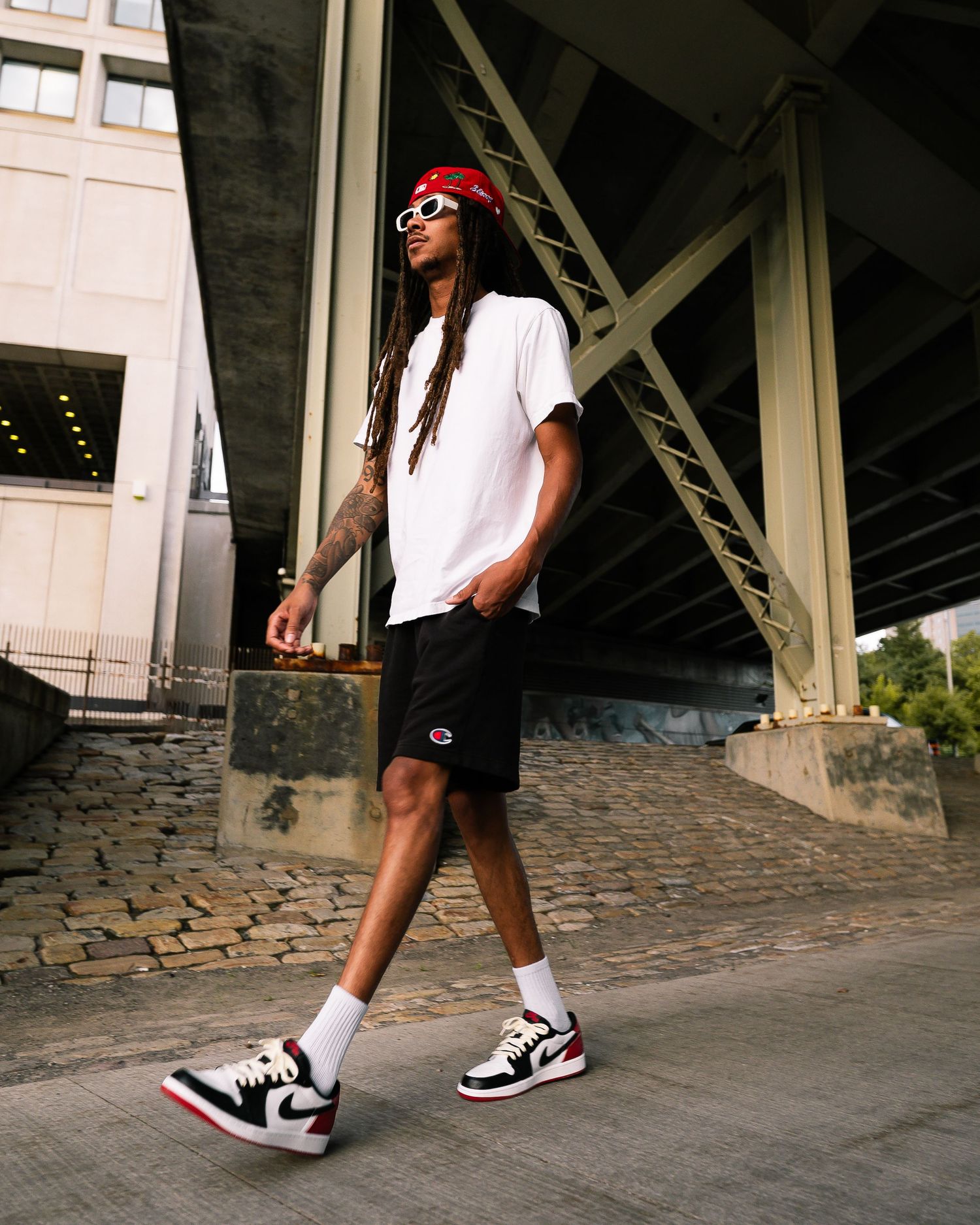 Person in white shirt and black shorts walking on cobblestone path under bridge.