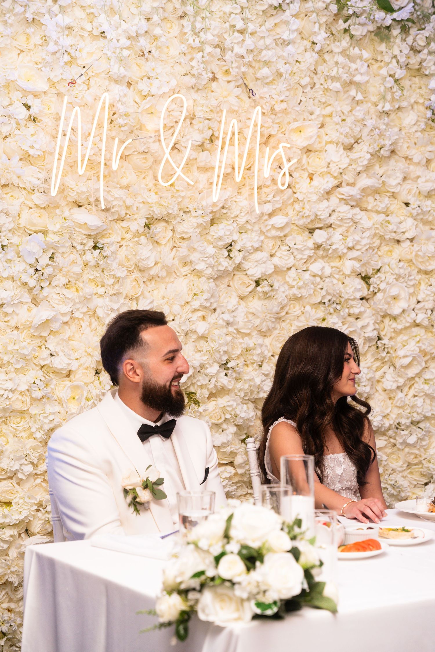 Wedding reception scene with a couple seated at a decorated head table with white floral arrangements.
