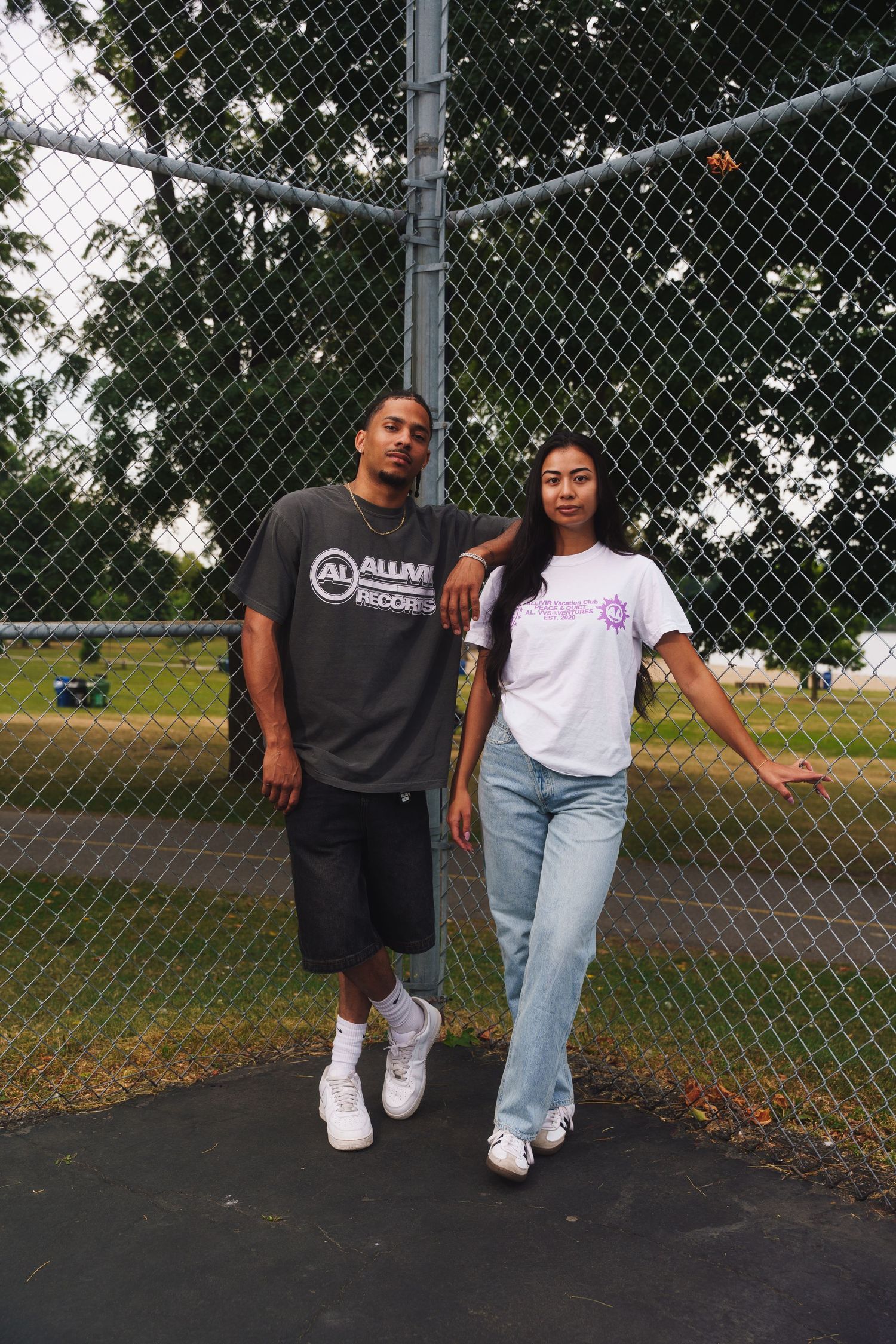 Two people in casual wear posing against chain link fence at tennis court.