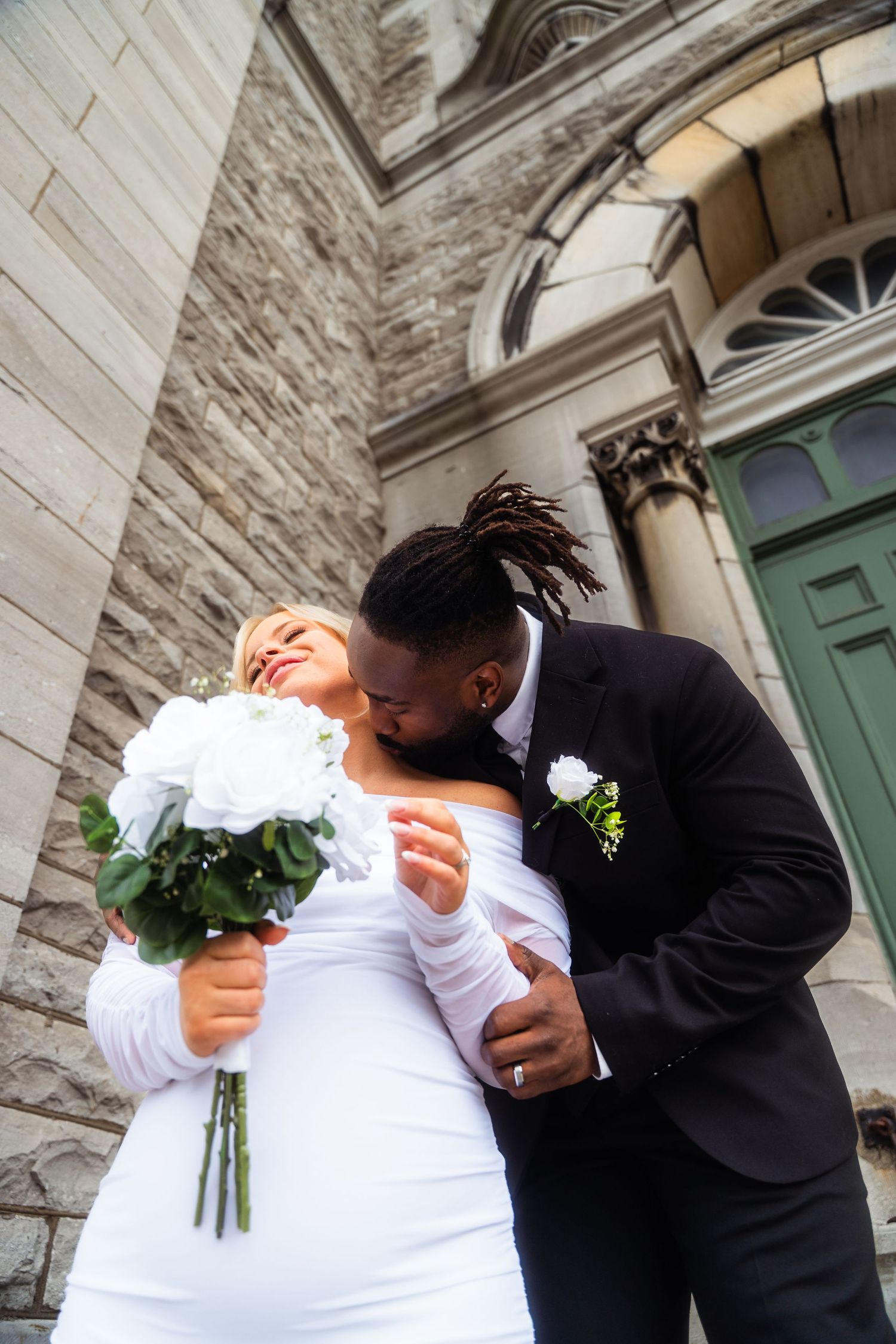 A joyful wedding couple embraces outside a historic stone church with an arched doorway and ornate architecture.
