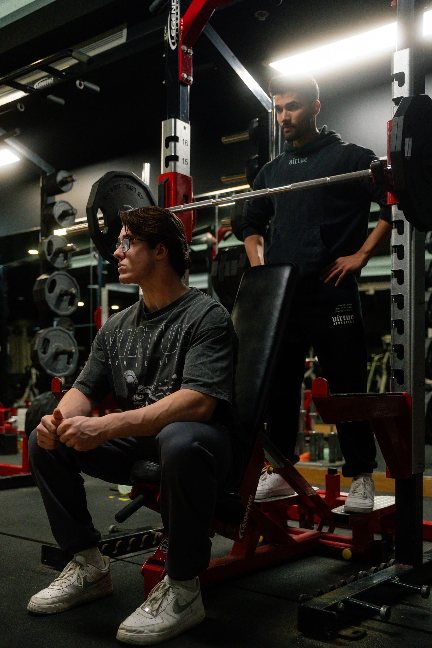 Person sitting on workout bench in dark gym wearing black clothing and white sneakers.