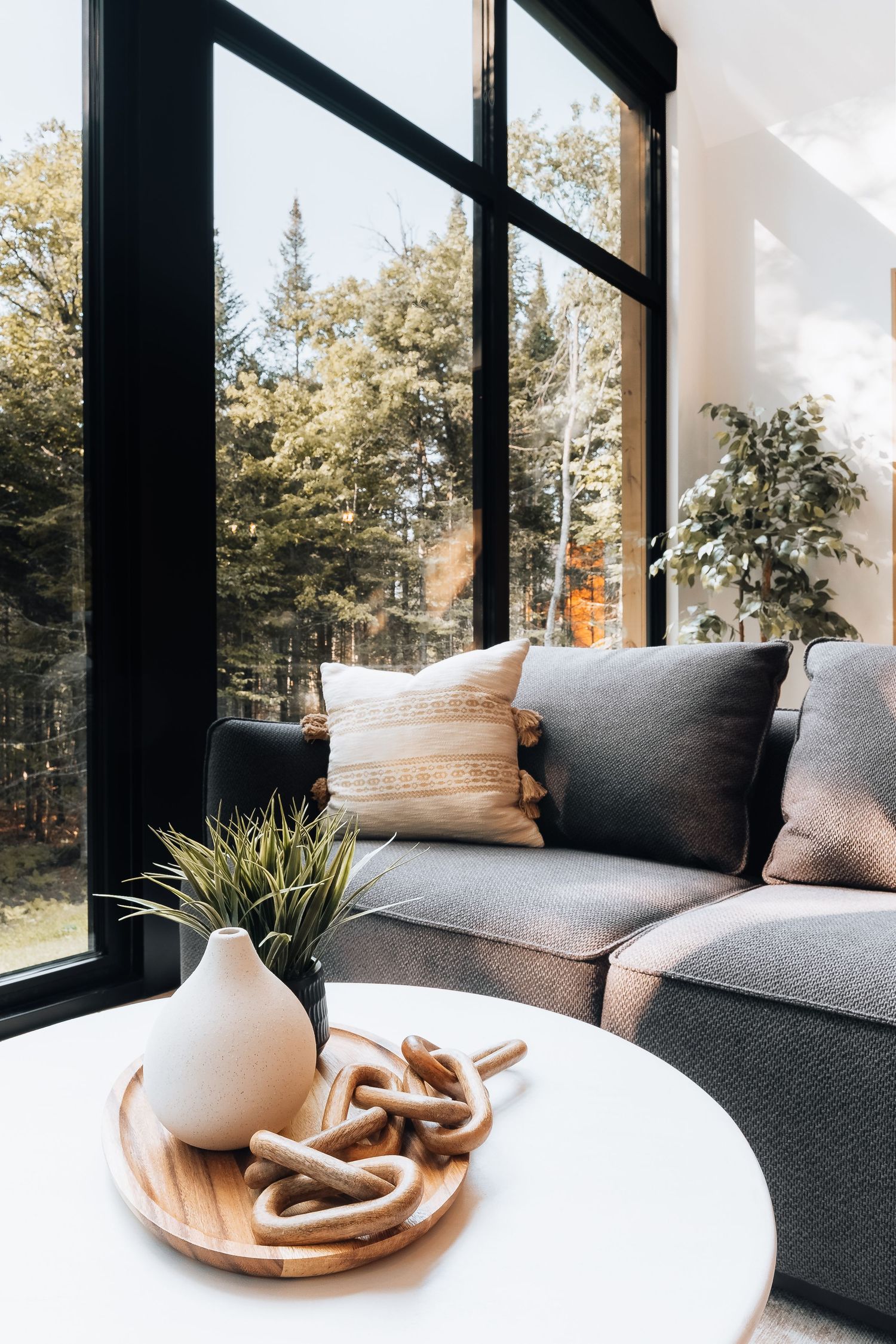 Modern living room corner with floor-to-ceiling windows, grey couch, and decorative elements on a round white table.