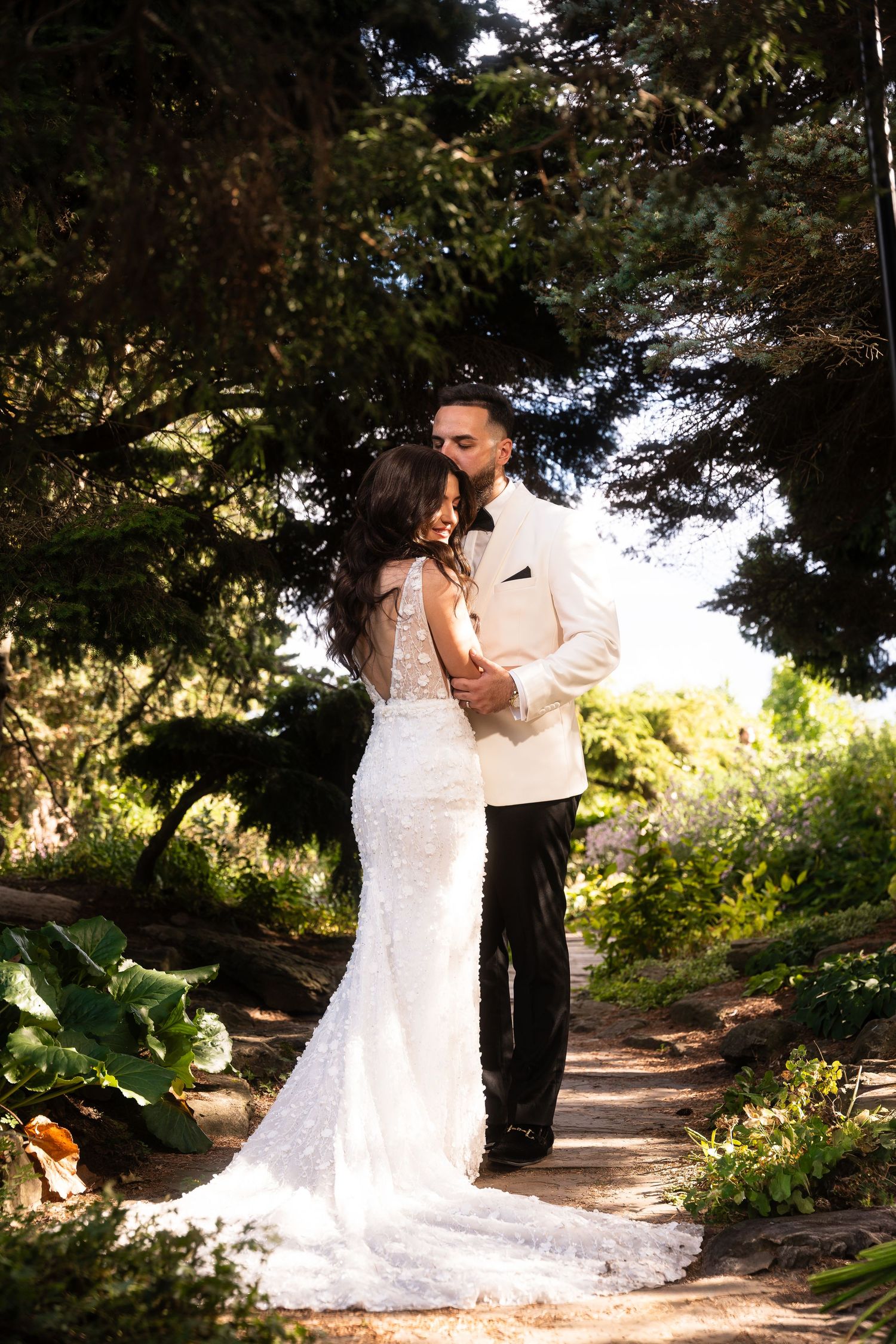 A couple in wedding attire dances together on a scenic garden path surrounded by lush greenery and trees.