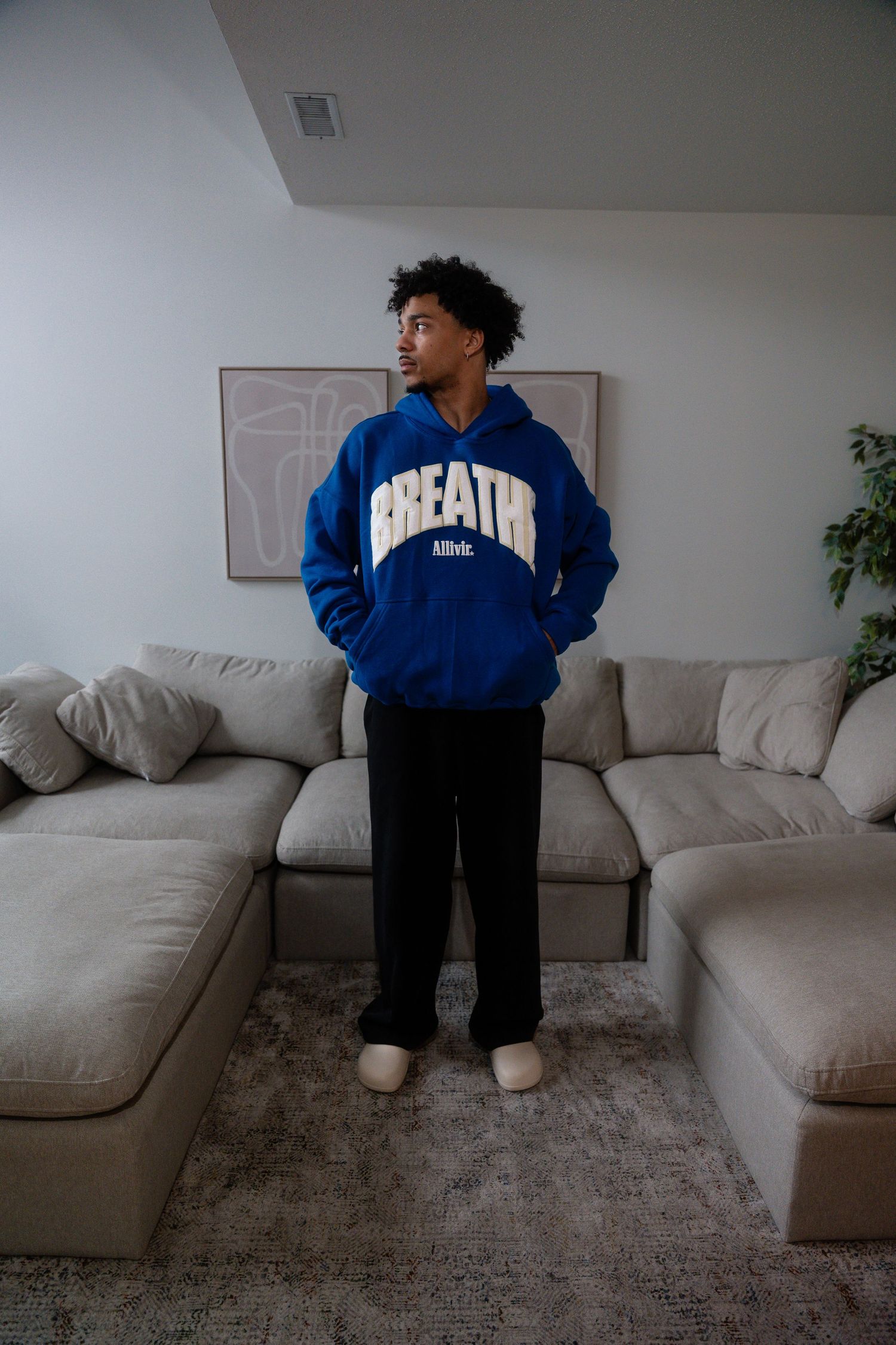 Person in a blue hoodie and black pants standing by a gray couch.
