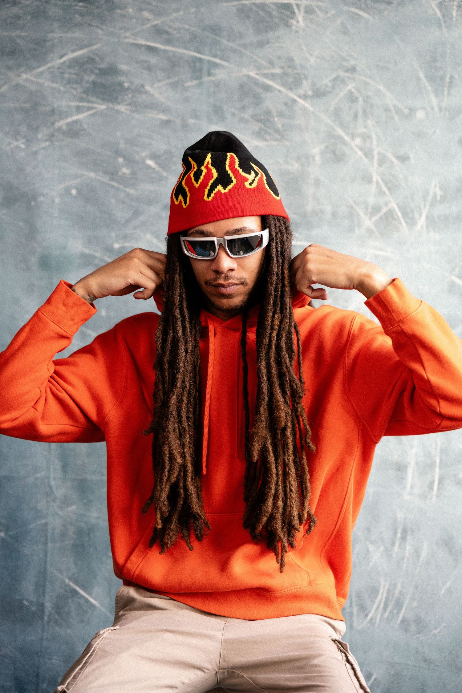 Person in orange hoodie and flame beanie striking poses against textured wall.