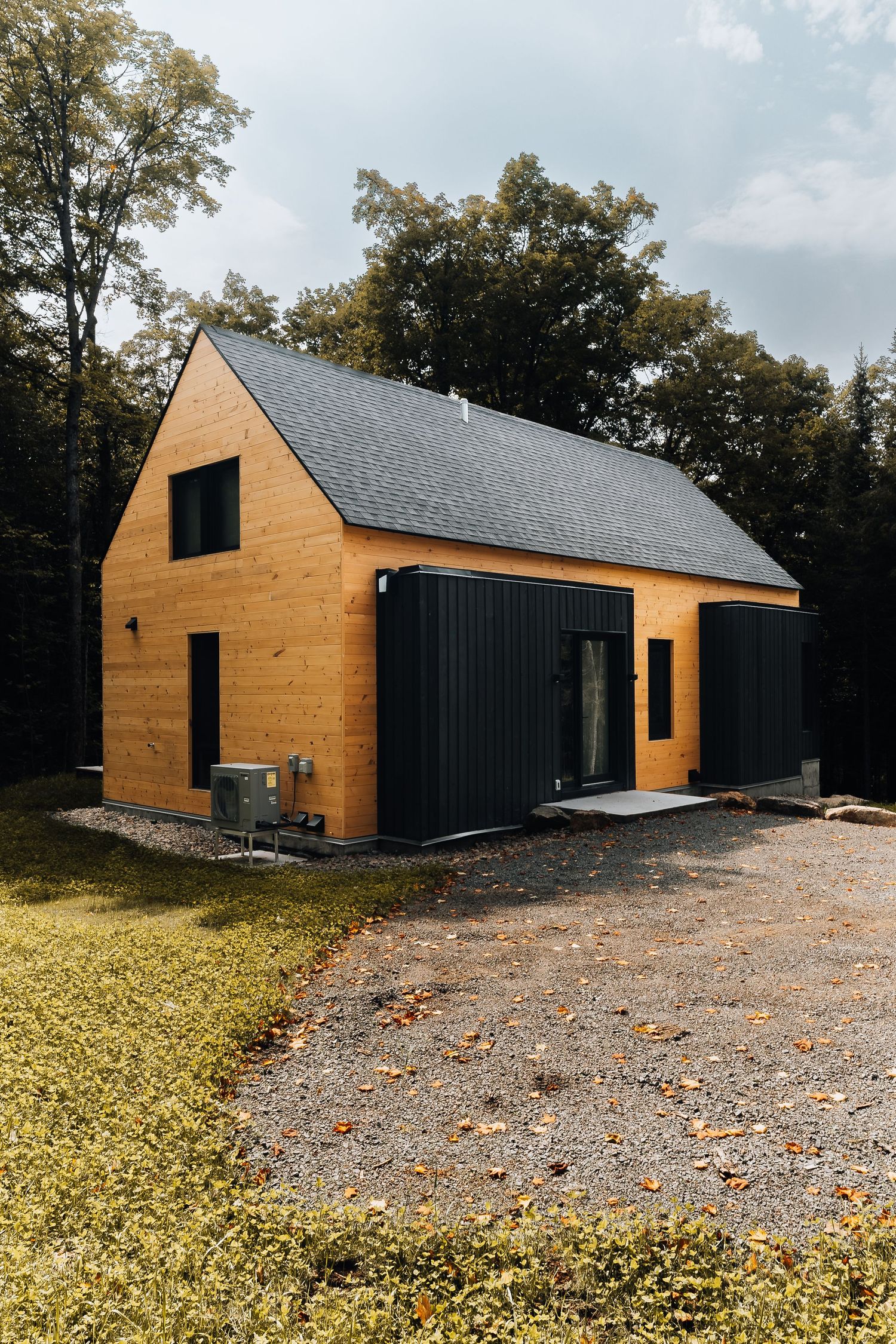 Modern wooden cabin with black accents and gravel driveway surrounded by autumn foliage.