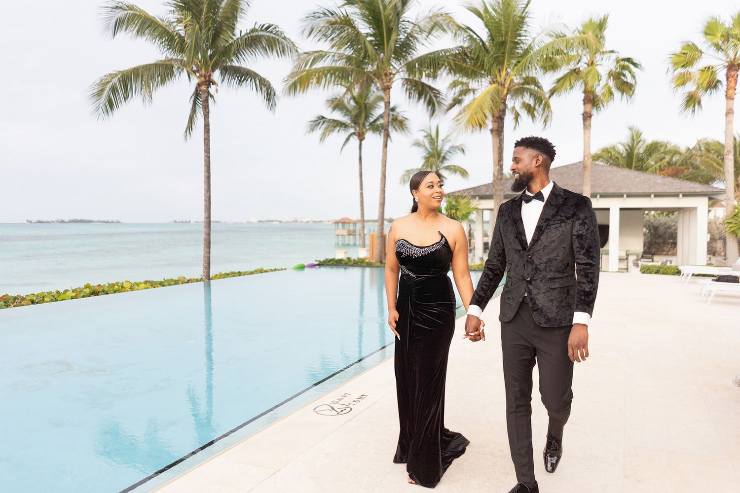 A couple in formal attire walks along an oceanside path with palm trees and turquoise waters in the background.