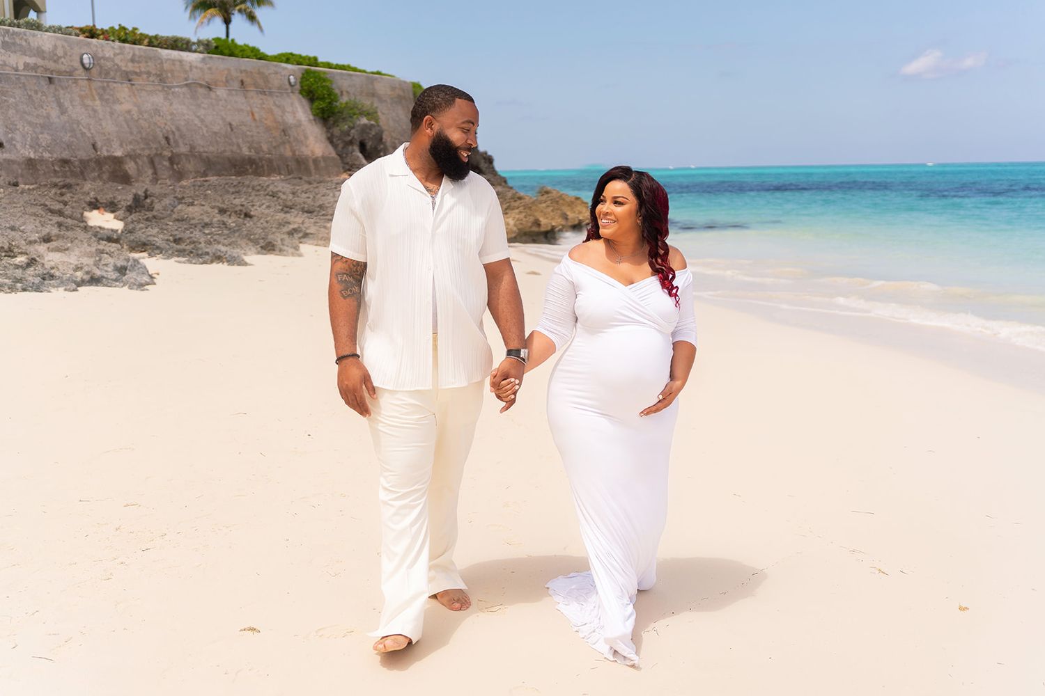 A couple in white outfits walking together on a tropical beach with turquoise water and cliffs in the background.